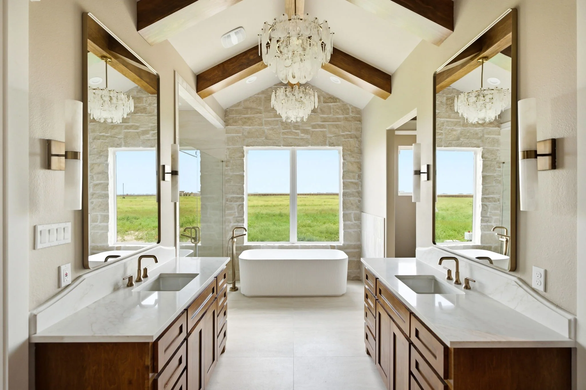 A bathroom with double vanities, white countertops, wooden cabinets, large wall mirrors, and a freestanding bathtub in front of a large window showing a green outdoor landscape; two chandeliers hanging from a vaulted ceiling with dark wooden beams.