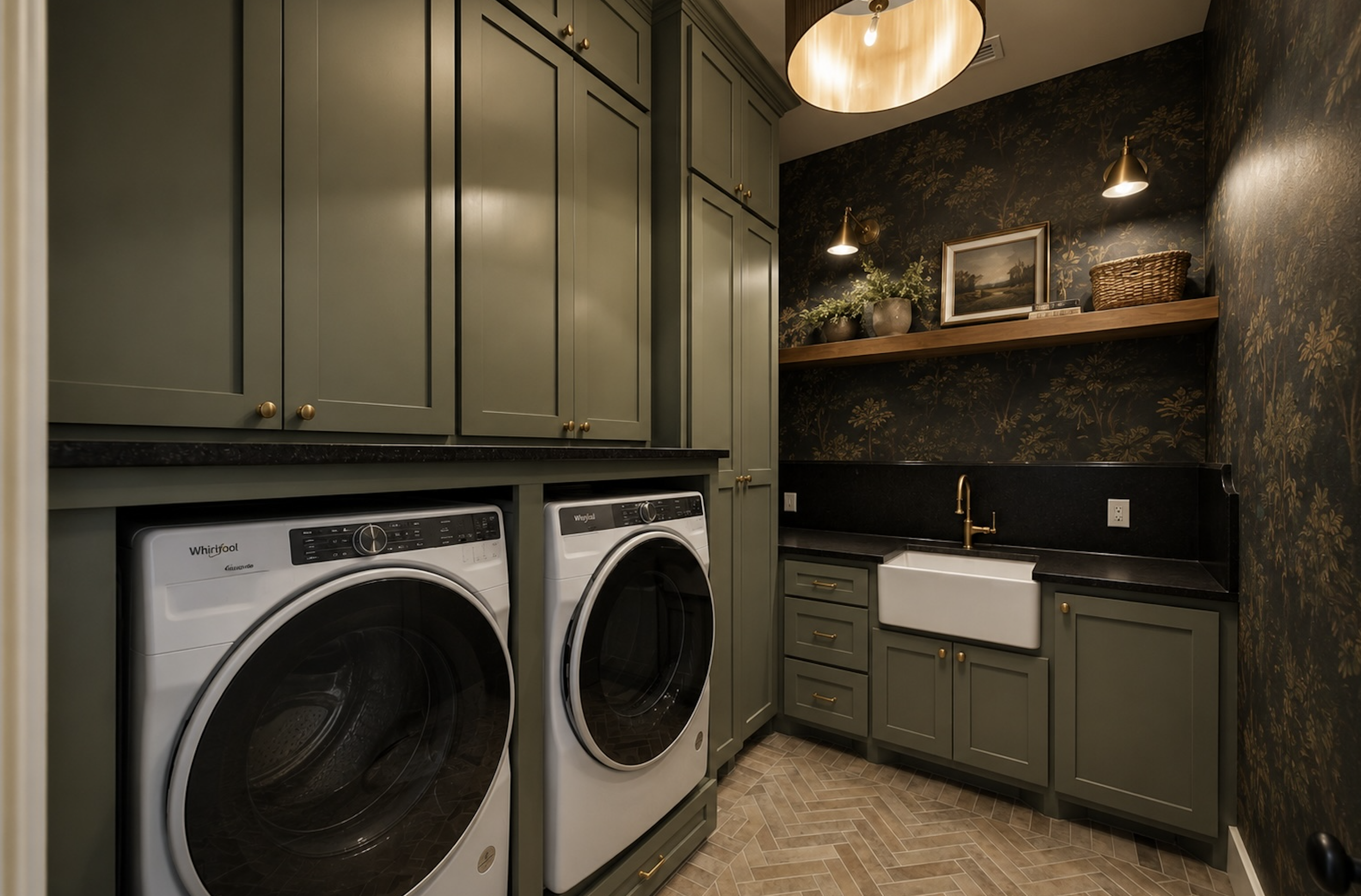 Laundry room with dark floral wallpaper, green cabinets with gold handles, white Whirlpool washer and dryer, wooden countertop with a white sink, gold faucet, decorative plant, framed landscape painting, and a woven basket on a wooden shelf.