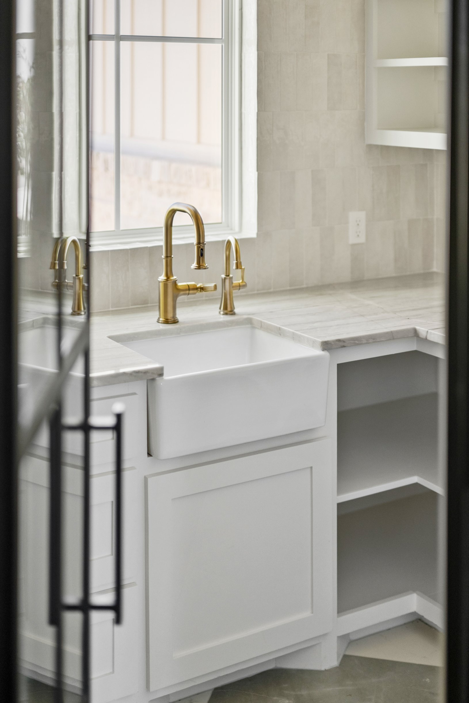 White kitchen sink with brass faucet in front of a window and open shelves on the wall.
