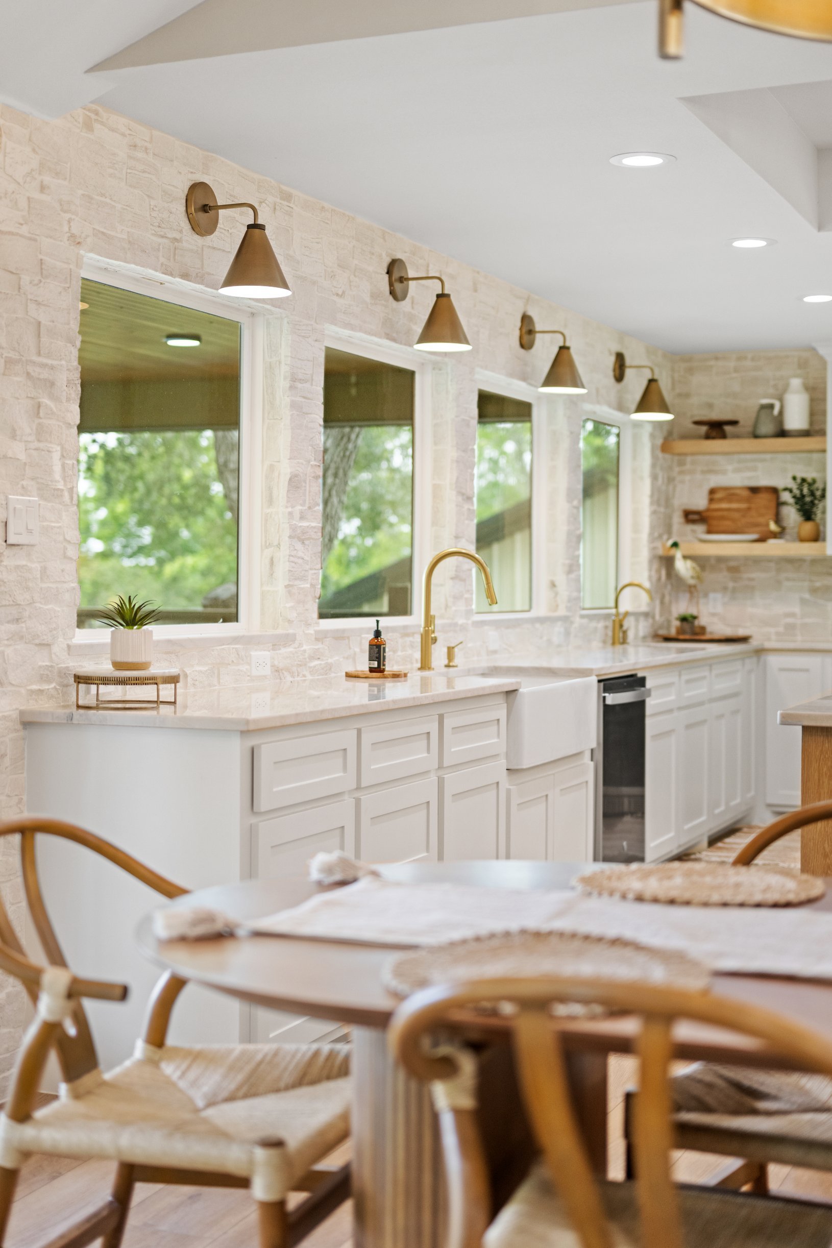 A bright kitchen with white cabinets, a marble countertop, gold fixtures, and large windows showing green trees outside.