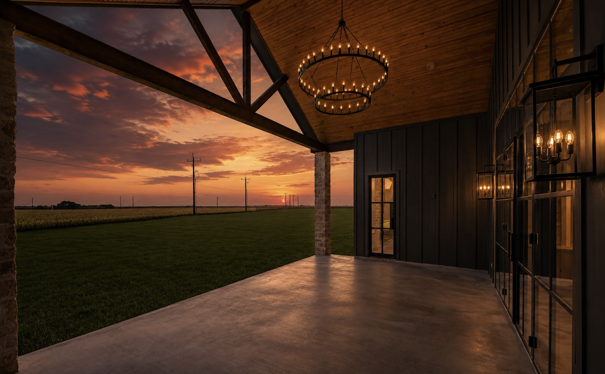A house patio at sunset with a wooden ceiling, black exterior wall, large glass door, and a circular chandelier with candles hanging from the ceiling. There is a field and power lines in the background.
