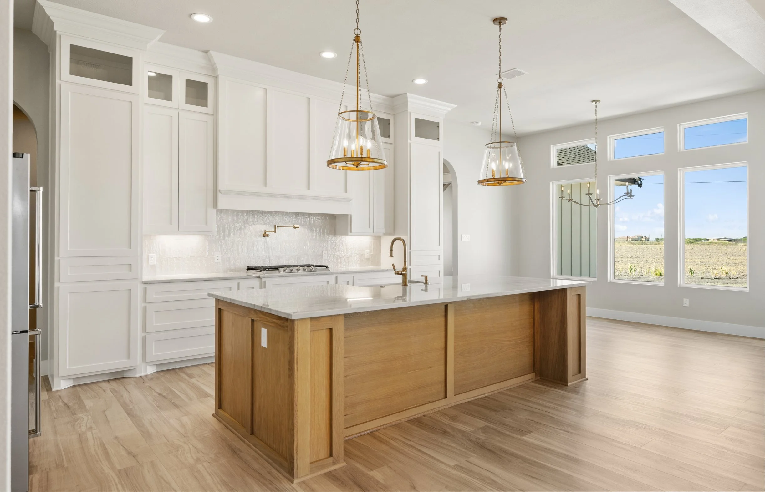 Modern kitchen with white cabinets, a central wooden island, pendant lights above the island, and large windows showing an outdoor landscape