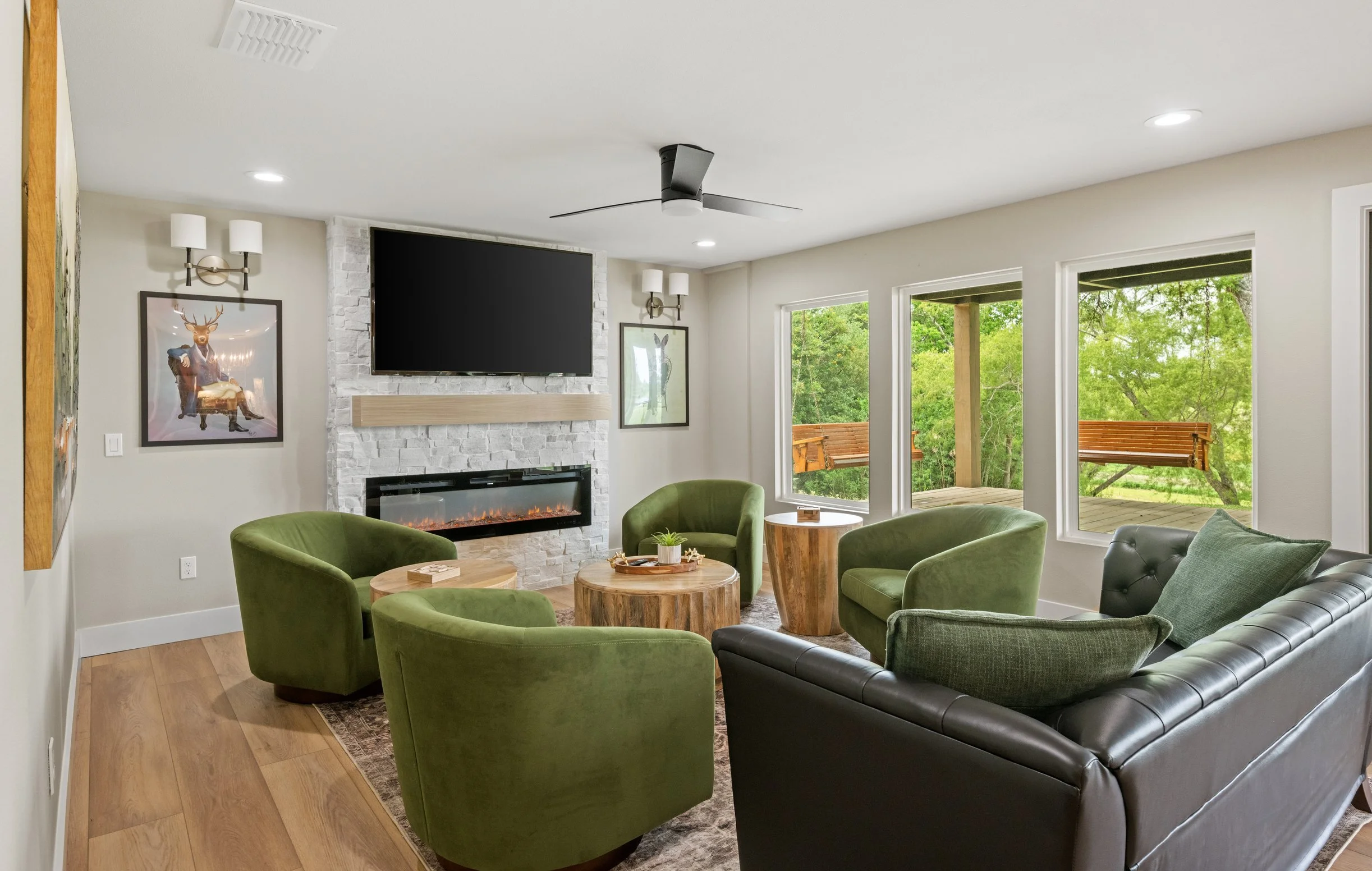 Cozy living room with four green armchairs, a black leather sofa, a wooden coffee table, a white brick fireplace, a large flat-screen TV, and windows overlooking a wooded outdoor area with a porch swing.