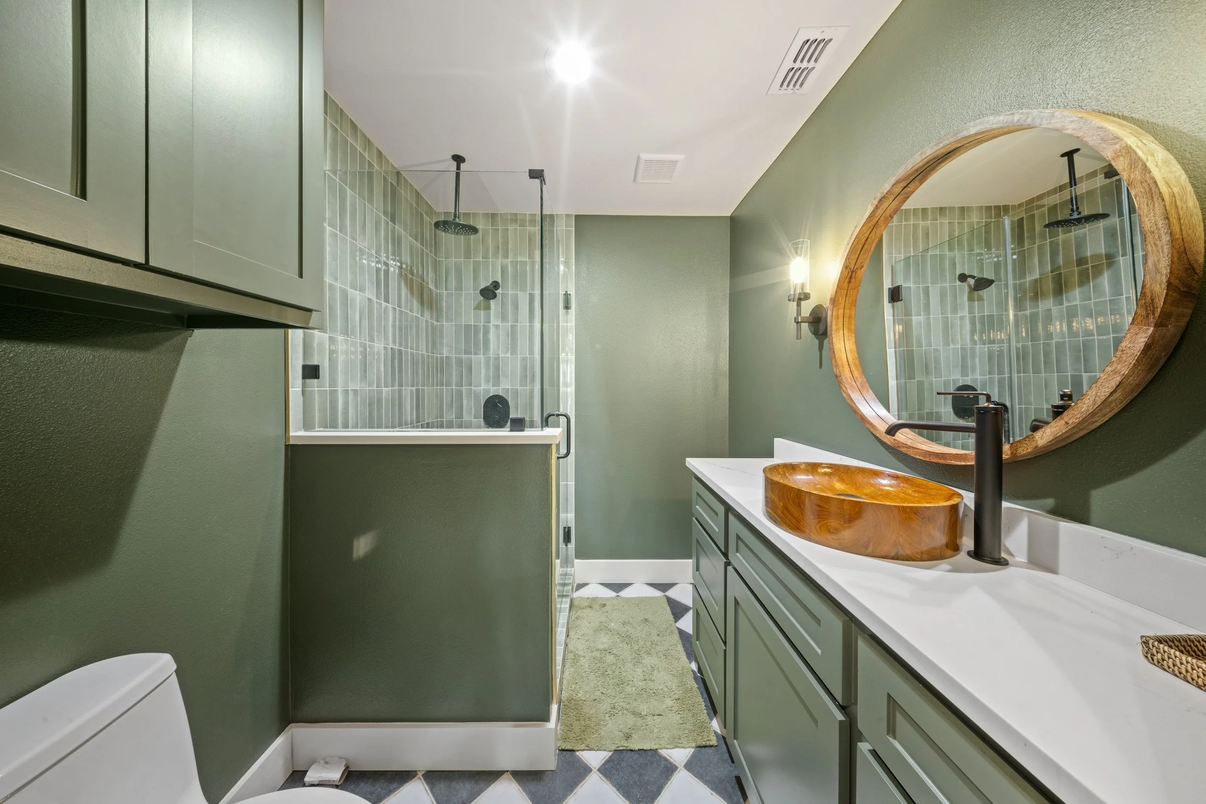 Bathroom interior with green walls, green cabinetry, a shower with green tile, a wooden sink vanity with a round wooden basin, a large mirror, and a small green rug.