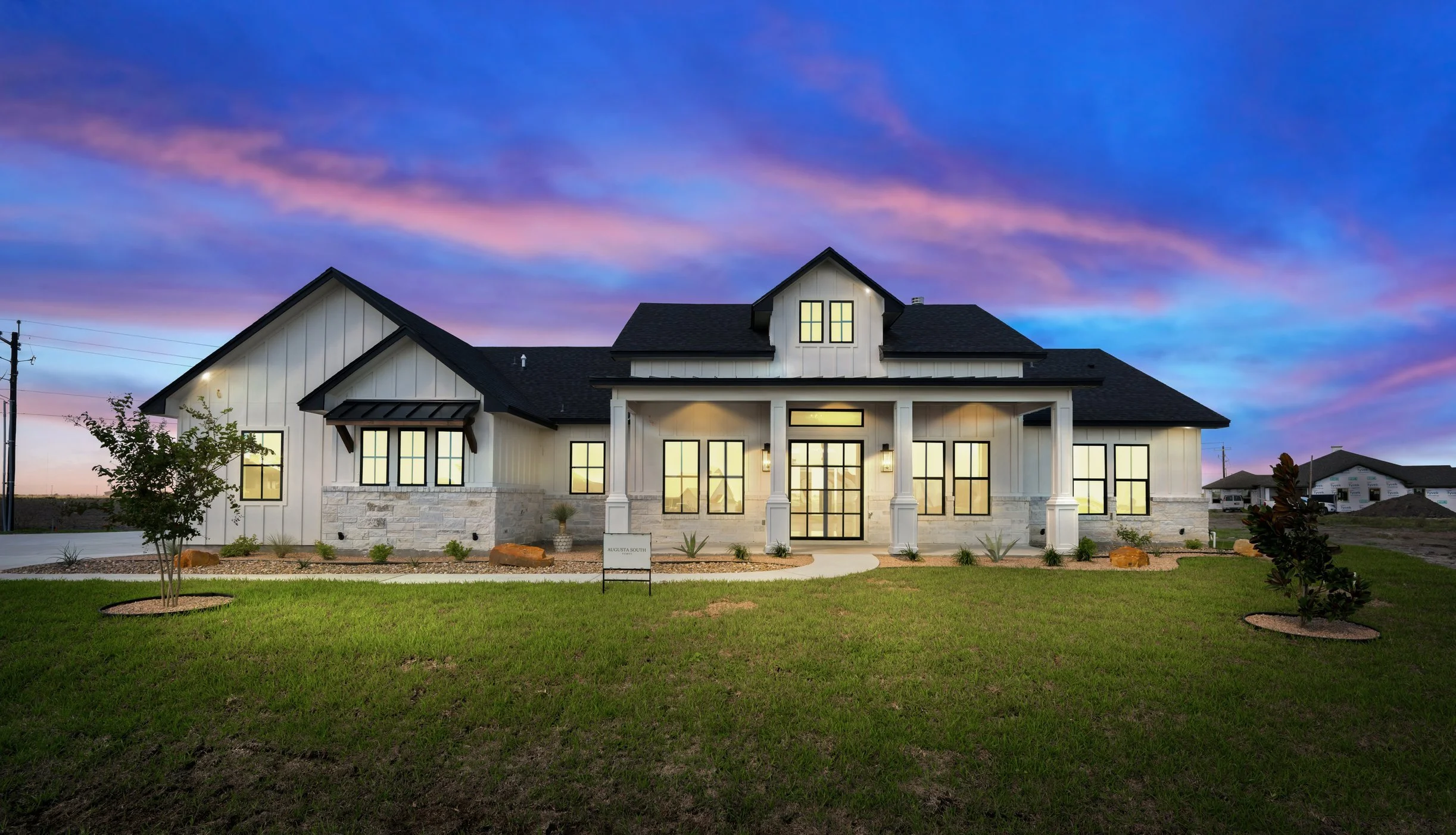 Modern two-story house with white siding and black roof, large windows, front porch with pillars, surrounded by a landscaped yard with small trees, at dusk with a colorful sky.
