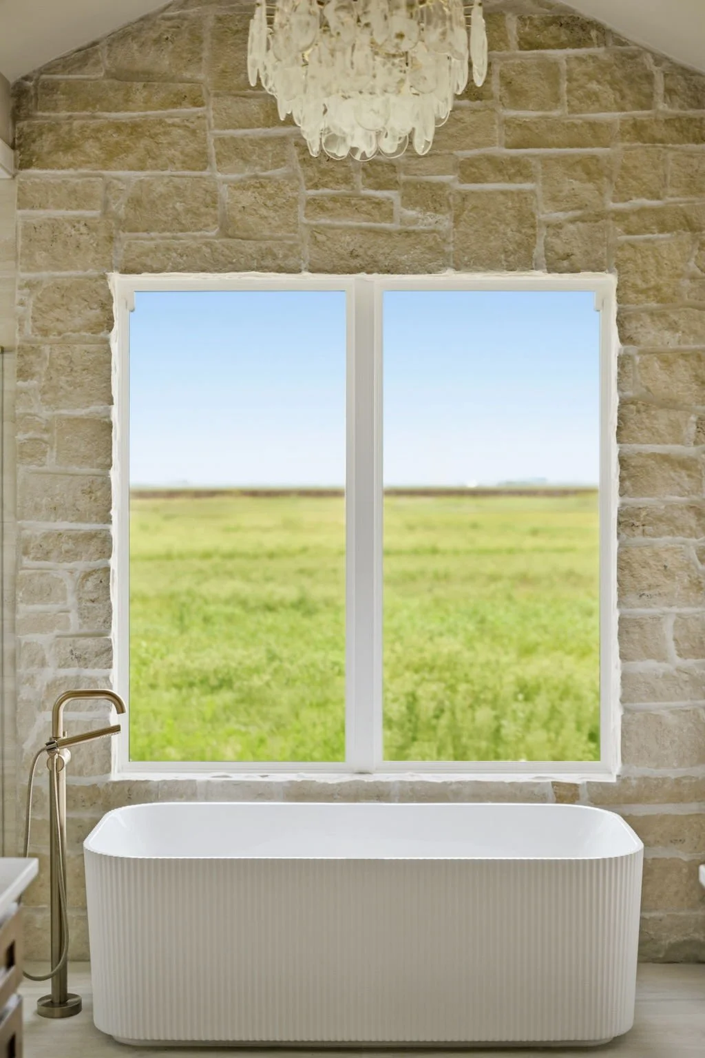 Bright bathroom with a white freestanding bathtub, stone wall, large window showing green fields and a clear blue sky, chandelier hanging from the ceiling.