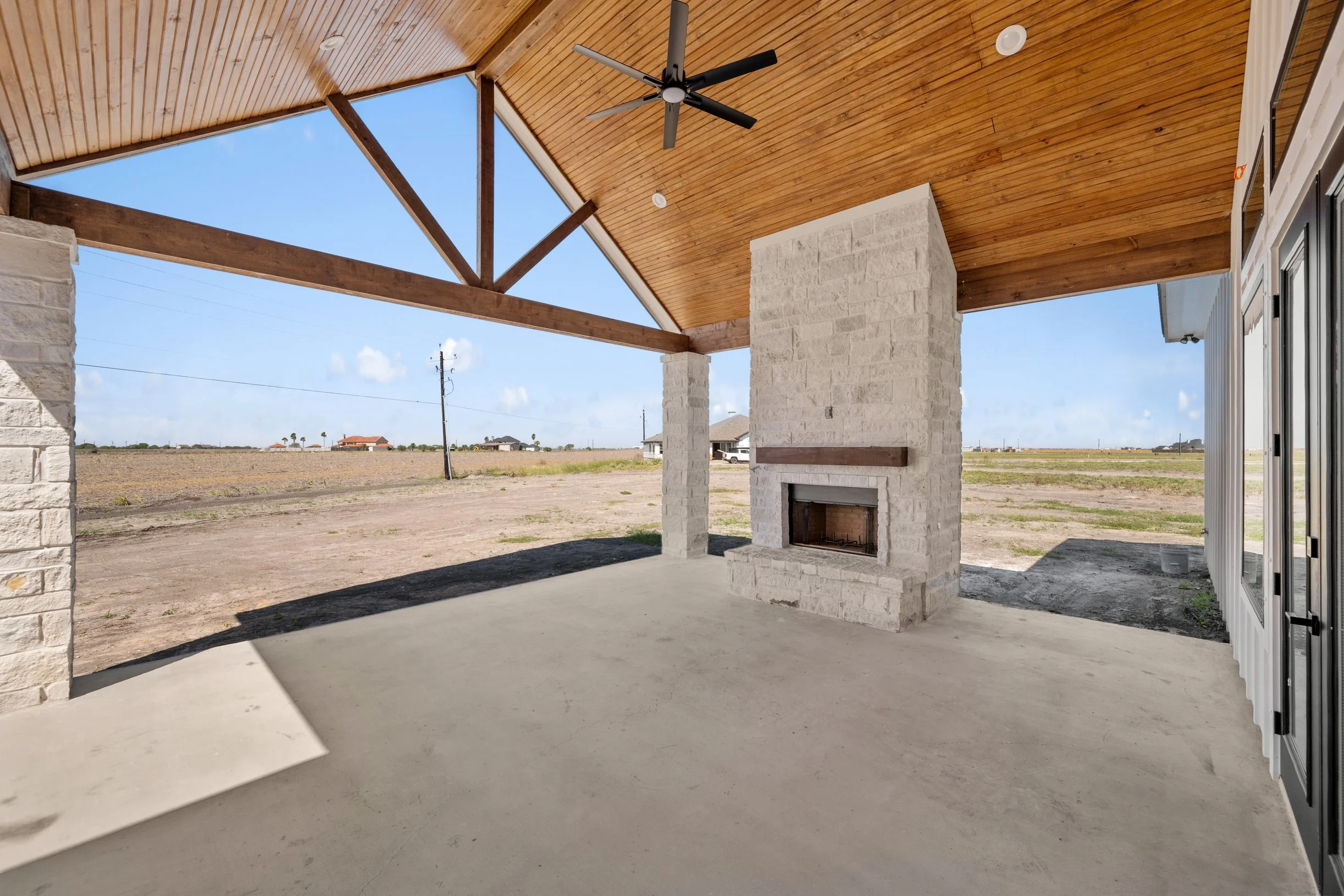 Covered patio area with a white brick fireplace, wooden ceiling with a ceiling fan, and open view of a rural landscape with a dirt field, utility poles, and distant houses.
