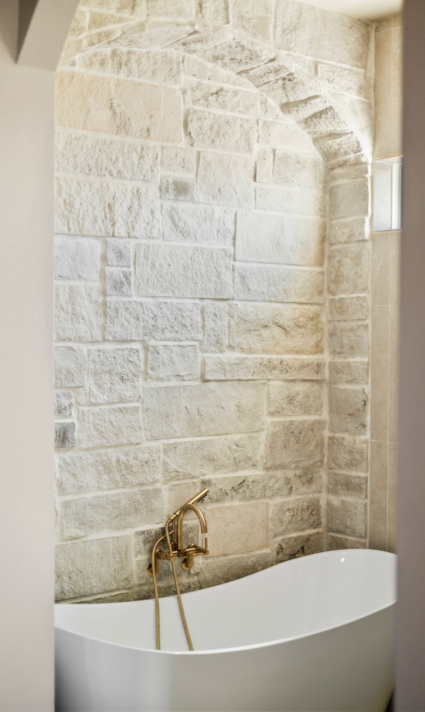 A modern bathroom with a white freestanding bathtub, a gold bathtub faucet with a handheld shower, and a textured light-colored stone wall.