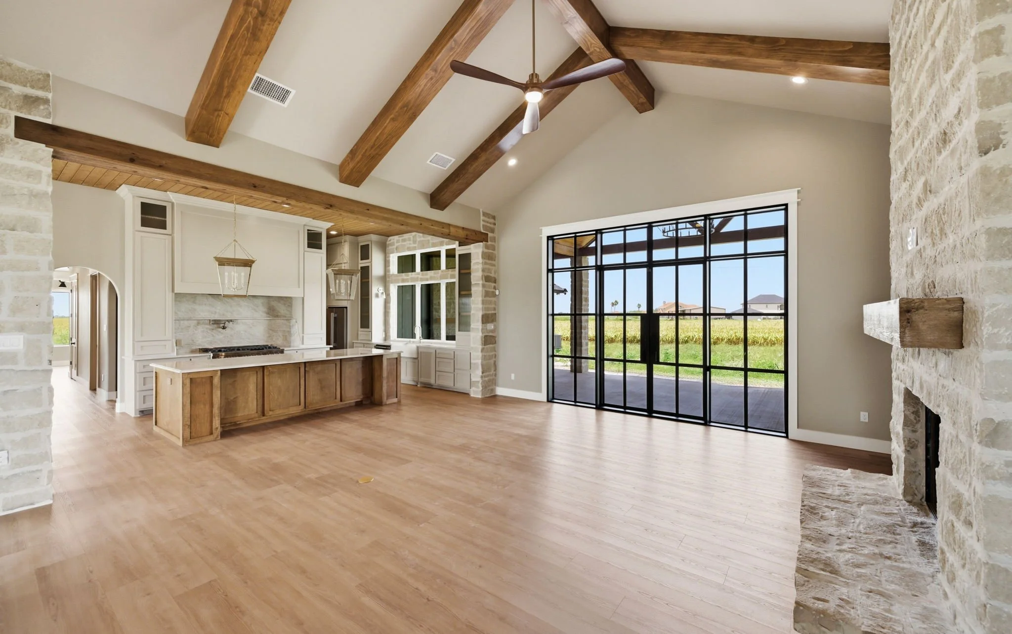 Open-concept living room and kitchen with high ceiling, exposed wooden beams, light-colored walls, large window, and sliding glass doors leading outside.