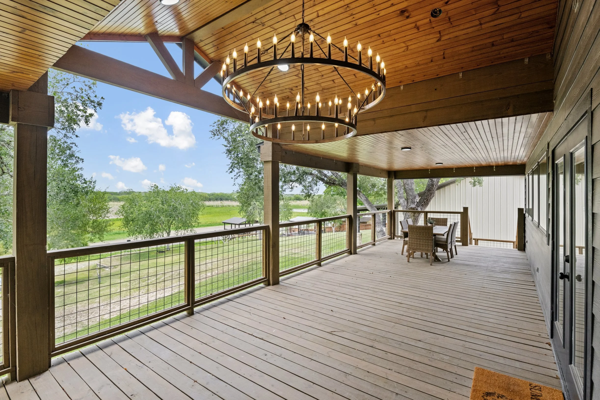 Covered porch with wooden ceiling and floor, outdoor dining table and chairs, railing, view of green trees and open sky, chandelier hanging from ceiling.