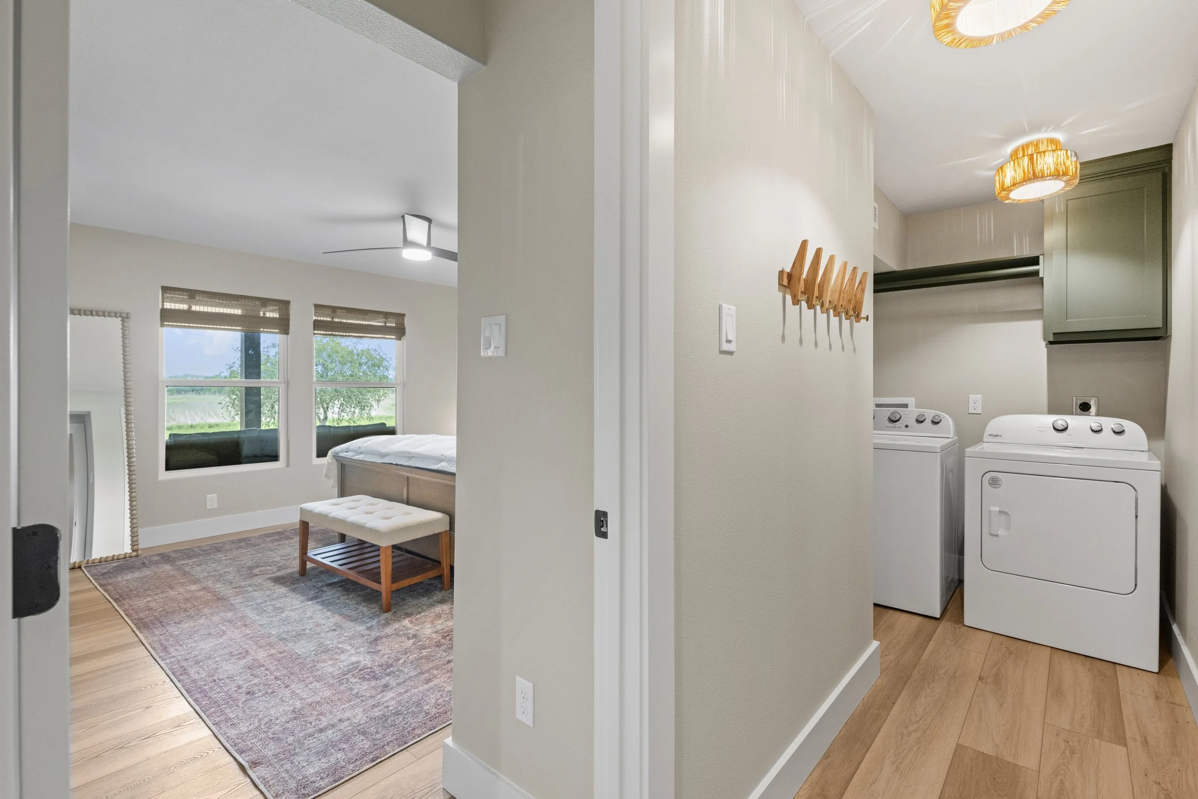 A bedroom with a window, a bed, a rug, and a bench, visible through a doorway. Next to the bedroom, a laundry area with a washer and dryer, green cabinetry, a wall-mounted coat rack, and ceiling lights.