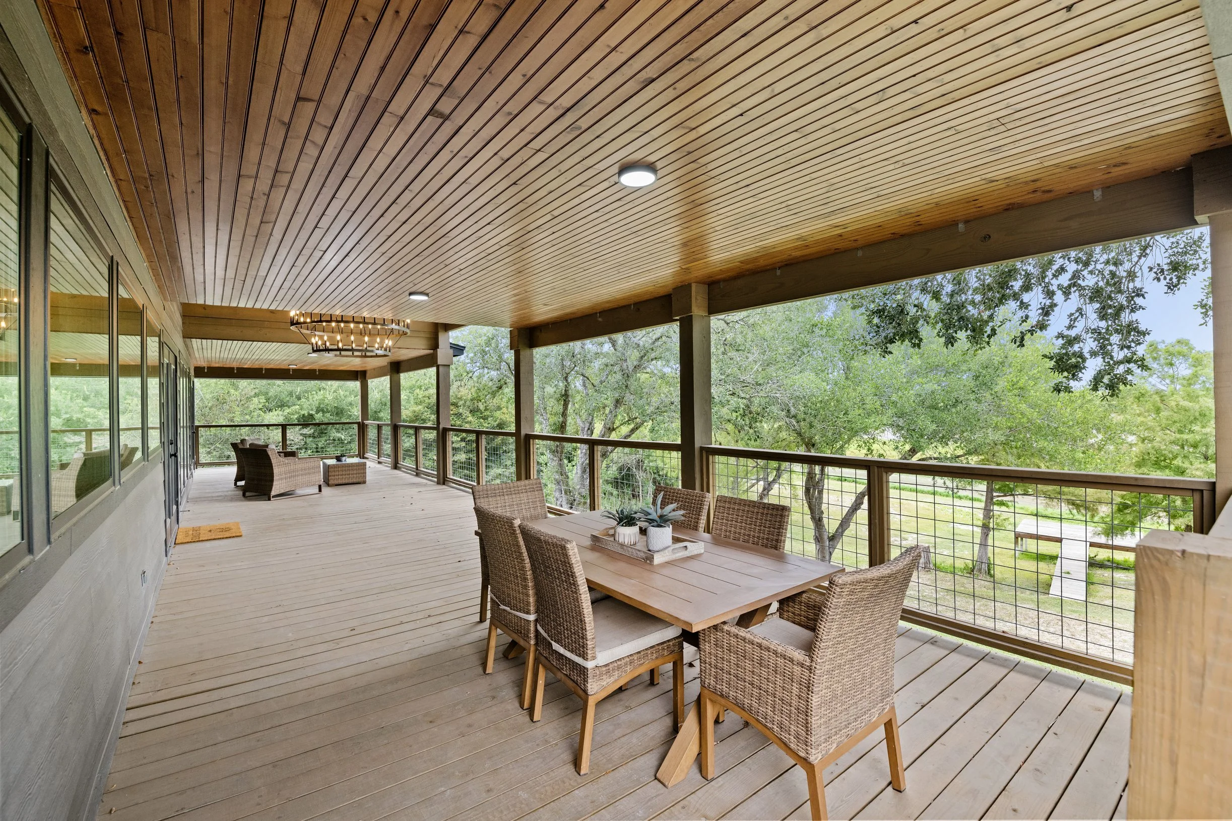 A spacious outdoor balcony with a wooden ceiling, surrounded by wooden railings, furnished with a dining table with wicker chairs, and overlooking green trees.