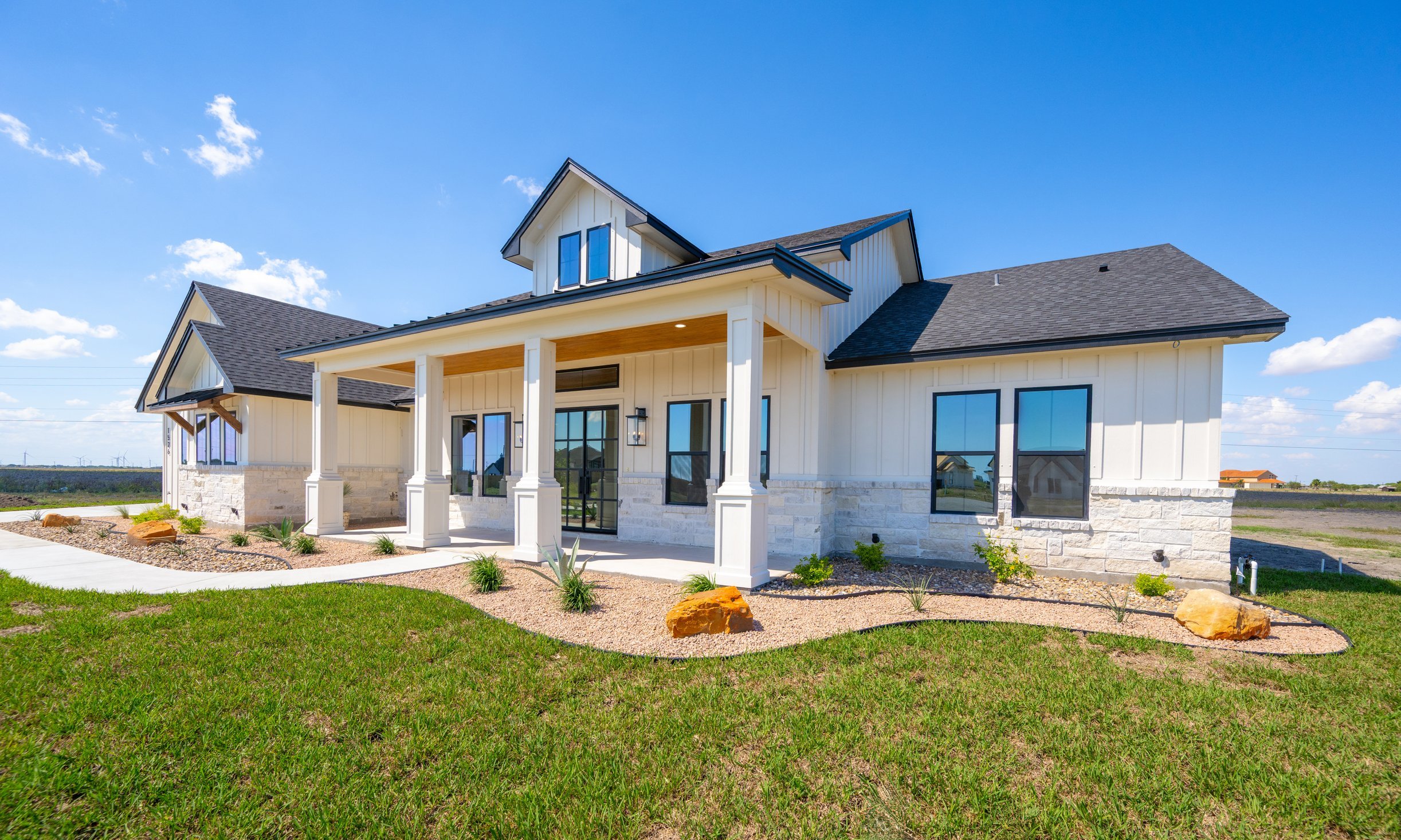 Modern house with white siding and black roof, front porch with white columns, large windows, surrounded by a landscaped yard with grass, rocks, and small plants, under a bright blue sky with a few clouds.