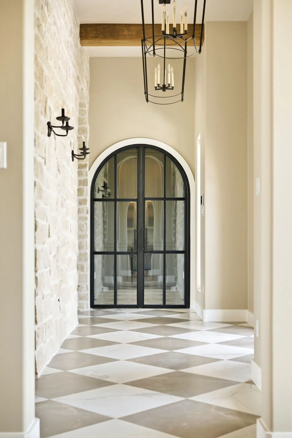 A hallway with a checkered floor, stone wall on the left, beige walls, and black metal chandelier and sconces. Double curved glass doors with black frames at the end.