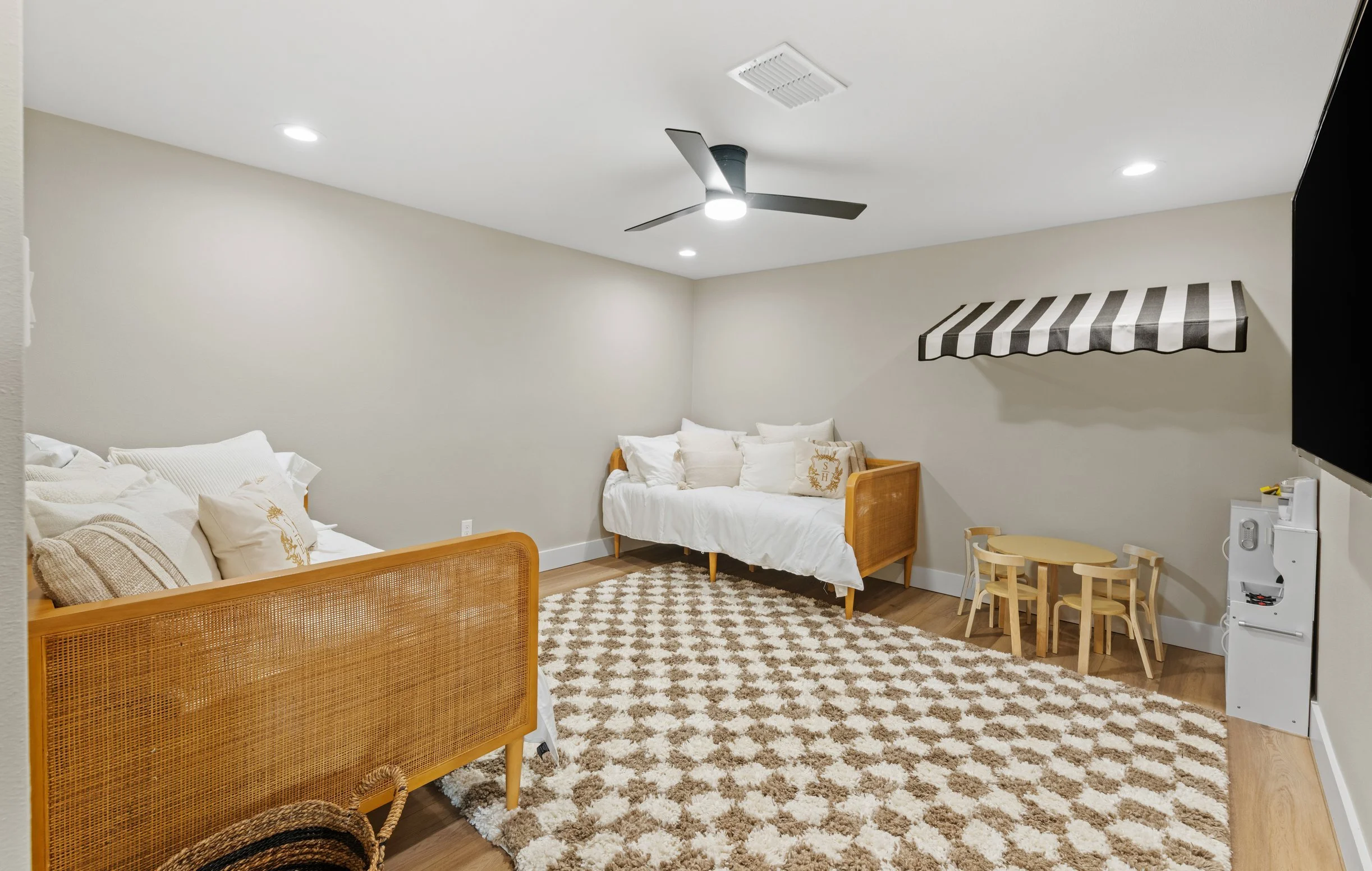 A cozy living space with beige walls and a patterned rug, featuring white sofas, small wooden chairs, and a striped awning on the wall. Recessed lighting and a ceiling fan are also visible.
