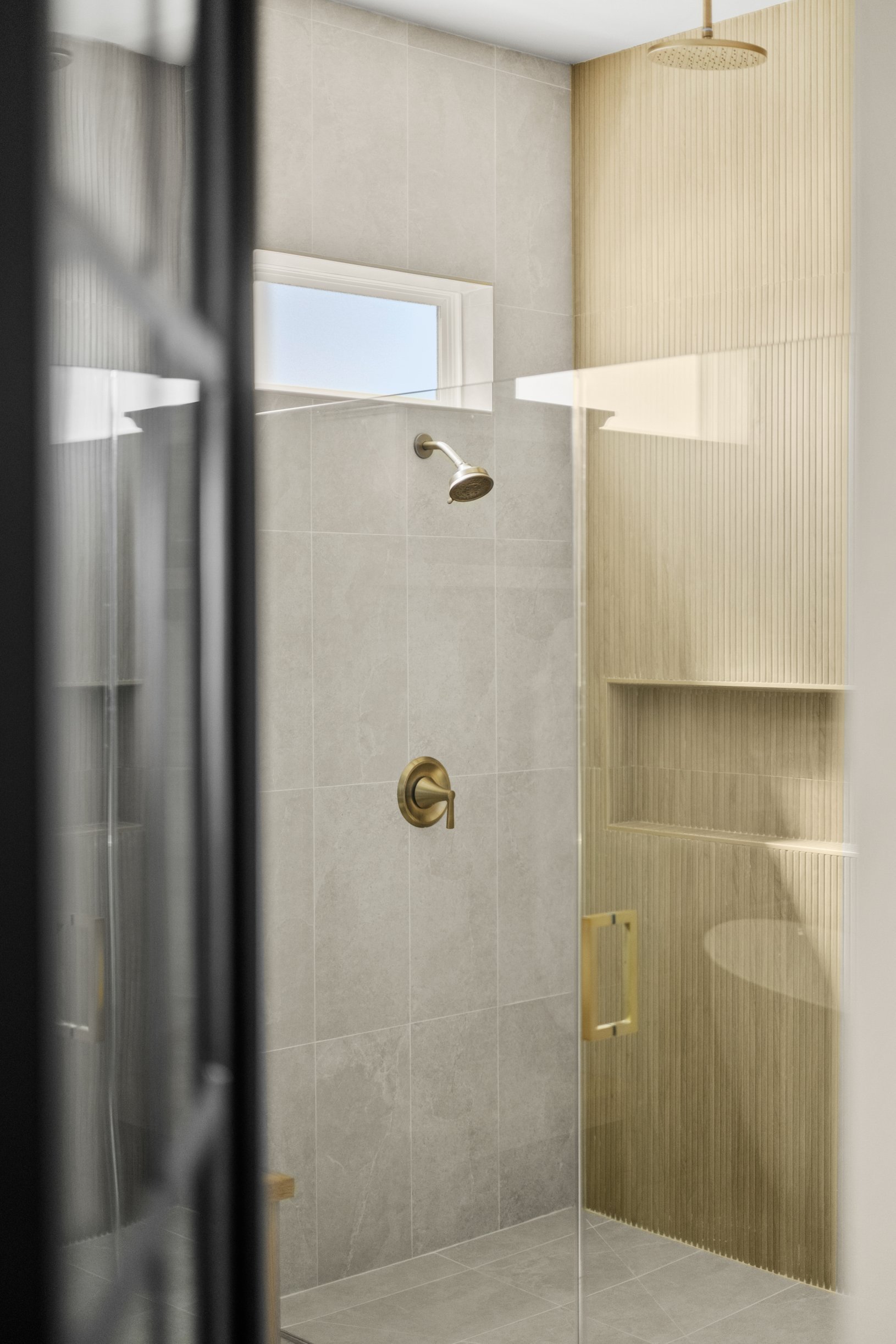 A modern shower with a rainfall shower head, a handheld shower head, and a glass door, featuring beige and gray tiles and a small window.
