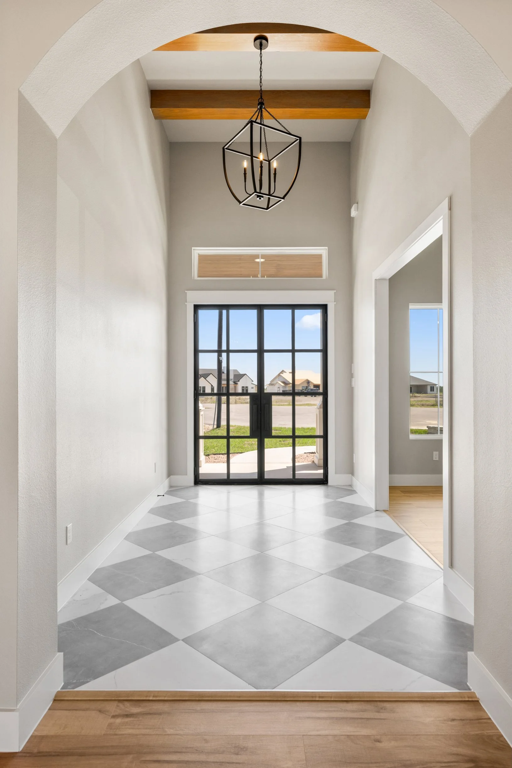 View through an arched doorway into a bright foyer with a large window, black-framed glass door, and a modern chandelier hanging from a ceiling with wood beams. The floor is patterned with white and gray tiles, and there is a transition to hardwood f