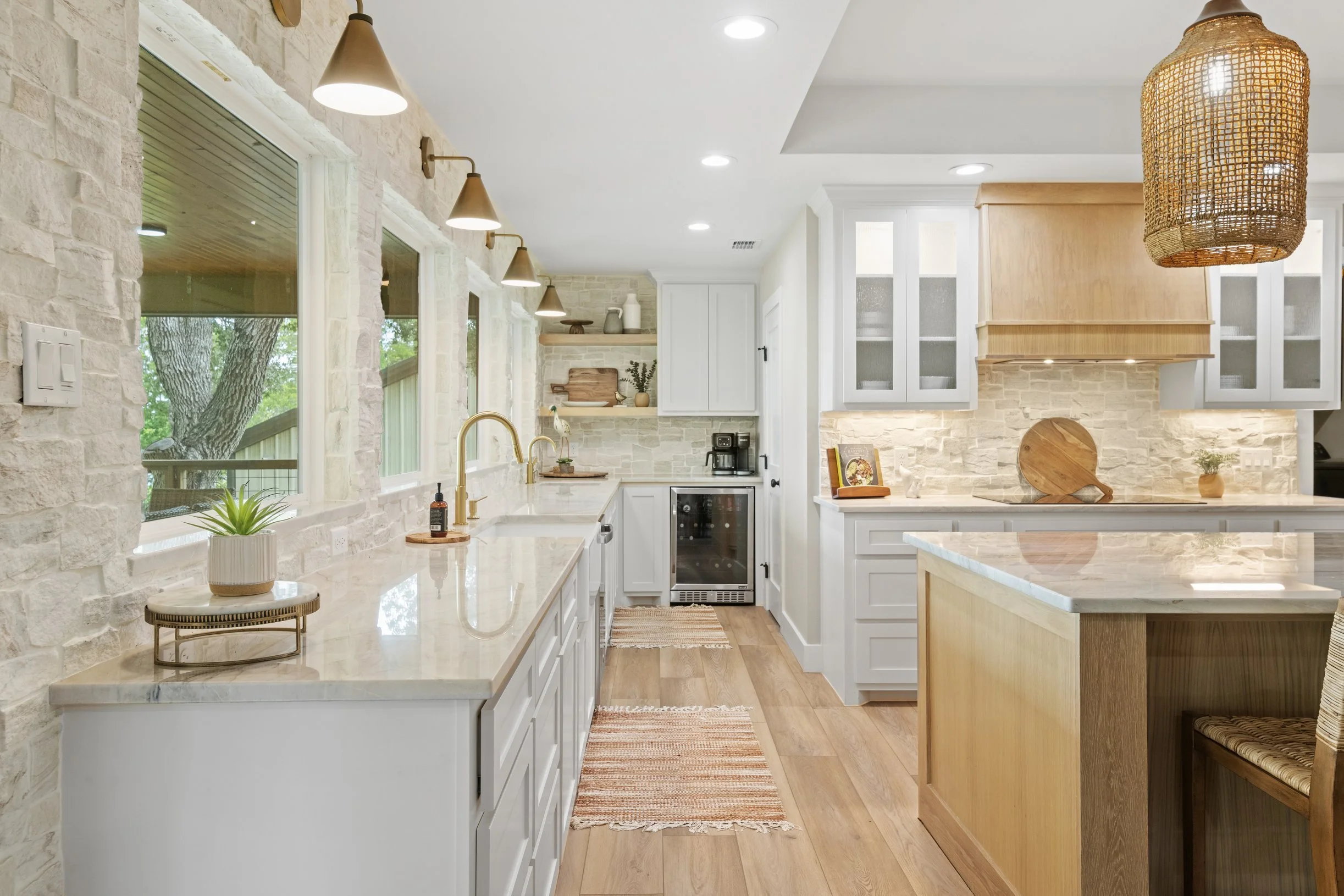Modern kitchen with white cabinets, stone backsplash, wooden accents, large windows, and warm lighting.