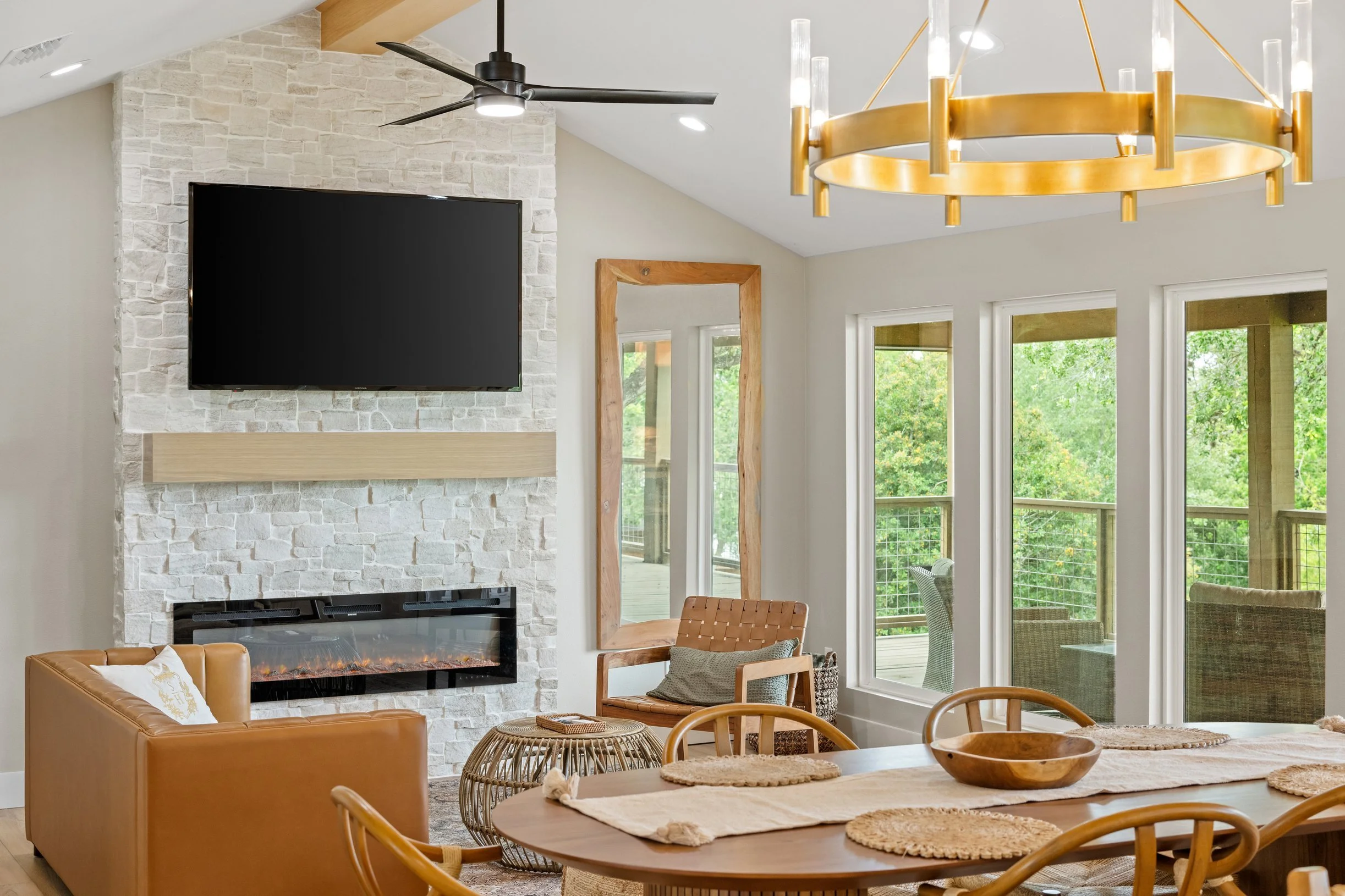 Living room area with a tan leather sofa, a round wooden dining table with woven placemats and a wooden bowl, a fireplace with a stone surround, a large flat-screen TV above the fireplace, a wooden frame mirror, a black ceiling fan, and a gold chande