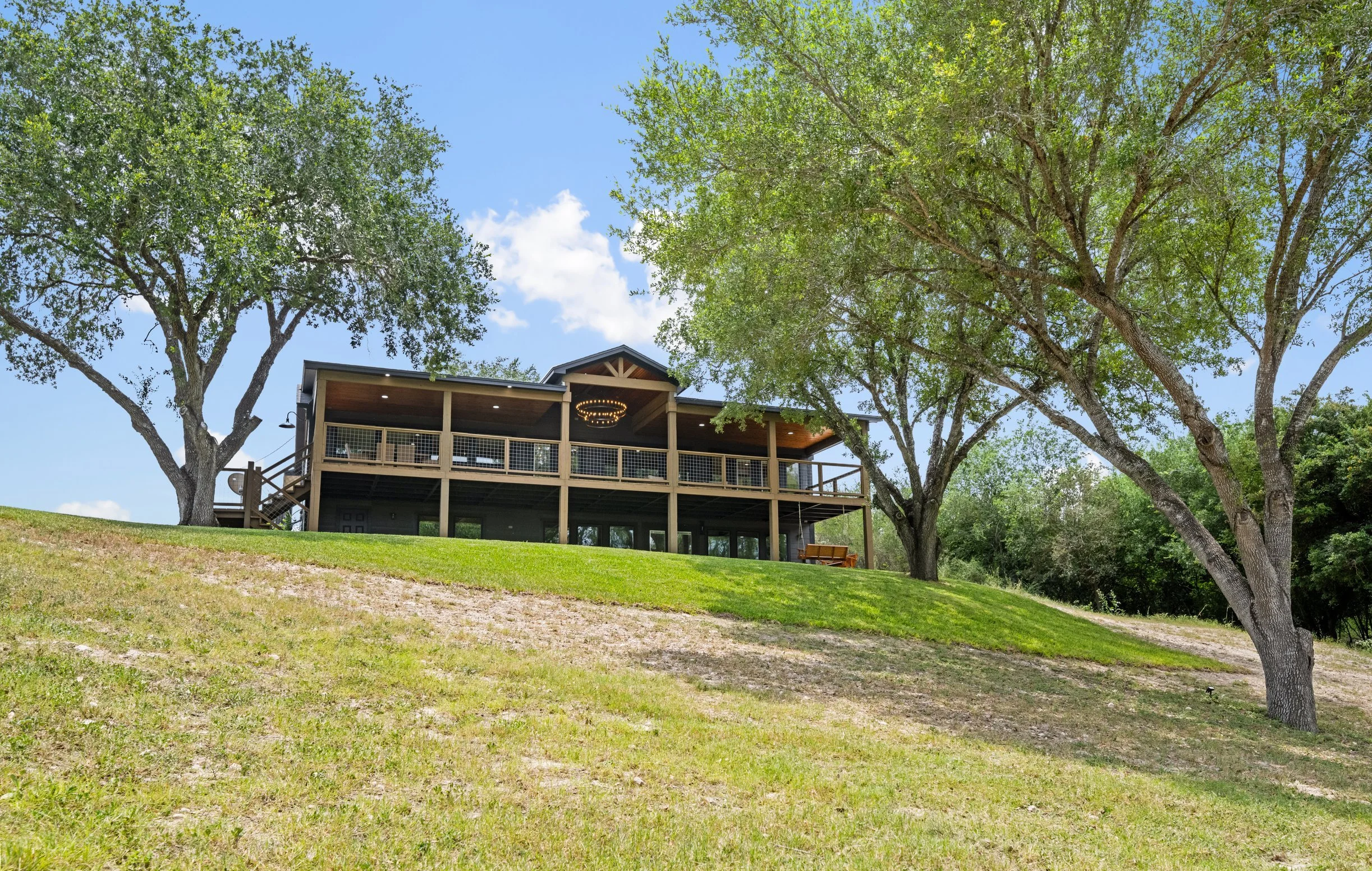 A two-story house with a wooden balcony, nestled among large green trees under a blue sky with some clouds.