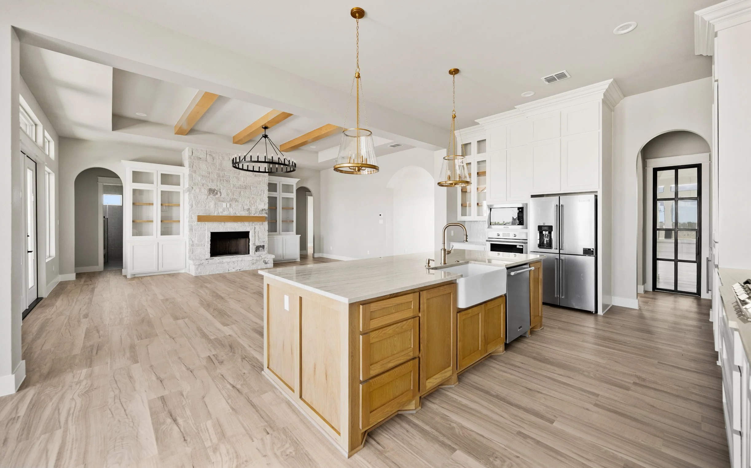Modern open-concept kitchen with light wood floors, white cabinets, a central island with a wooden base, stainless steel appliances, and a white brick fireplace in the background. Pendant lights hang above the island, and the ceiling features wooden 