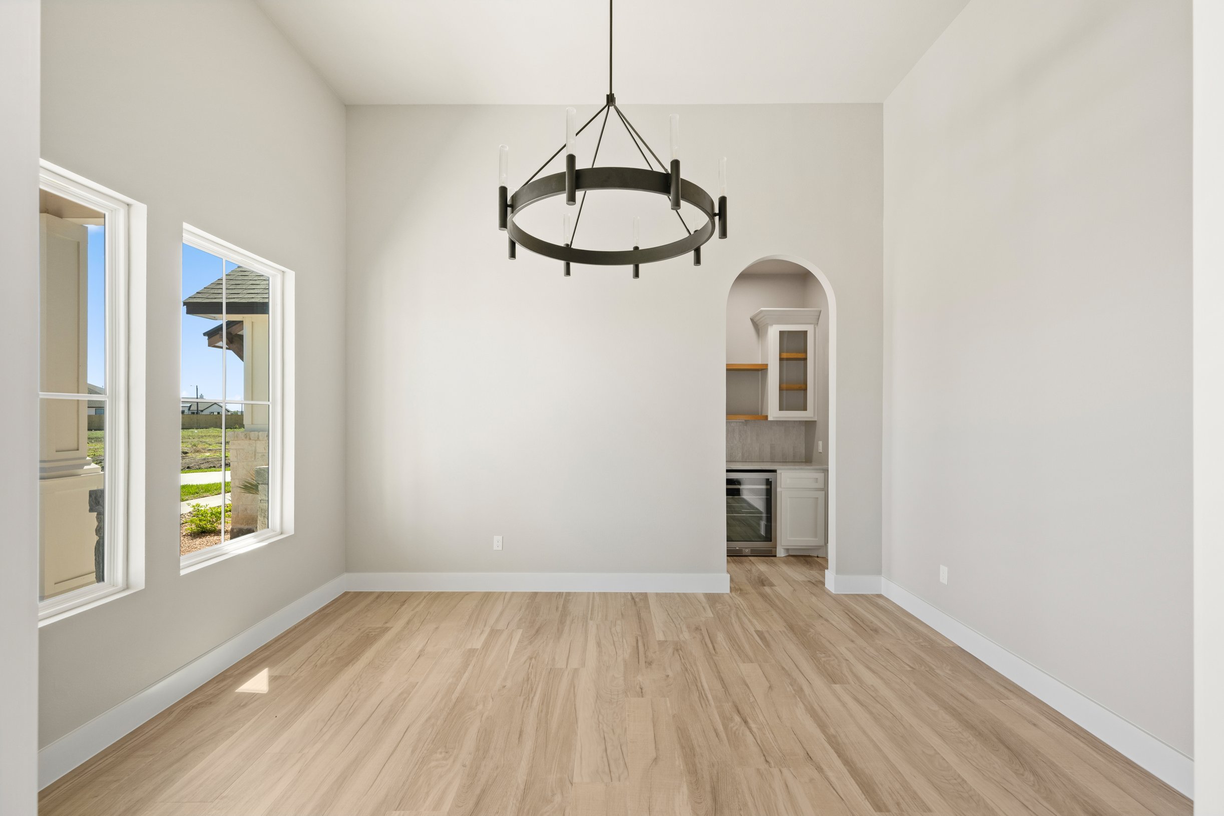 Empty room with light-colored wooden flooring, white walls, two large windows, and a black chandelier hanging from the ceiling. A small archway leads to a kitchen with white cabinets and built-in appliances.