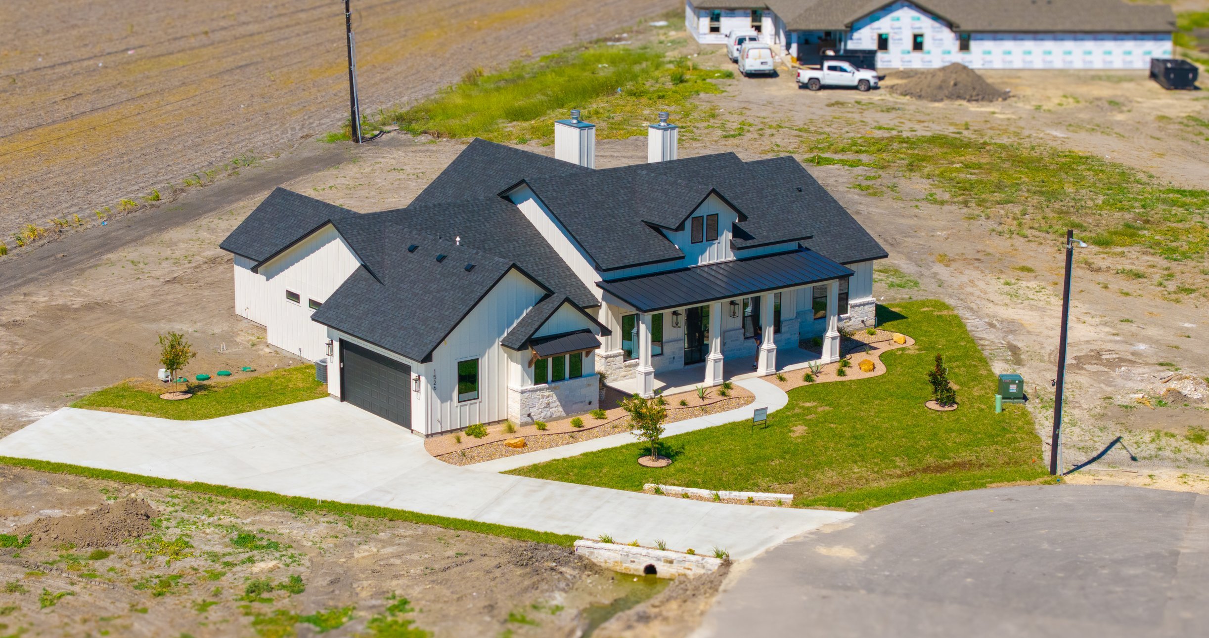 An aerial view of a modern house under construction with a finished front yard, driveway, and a partially built structure in the background.