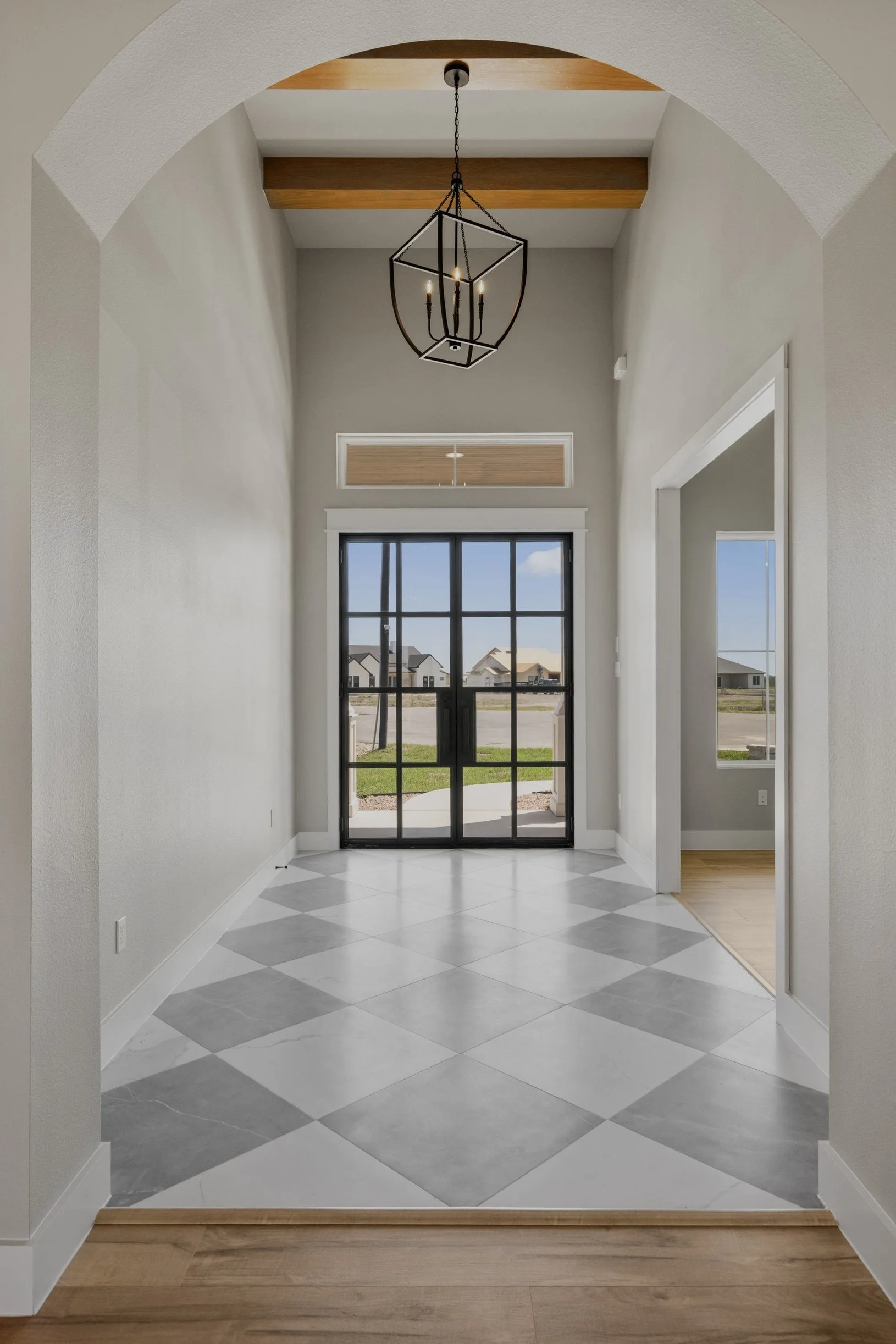 View through an arched doorway into a bright entryway with a large window, tile floor, and a modern chandelier hanging from a wooden ceiling beam.