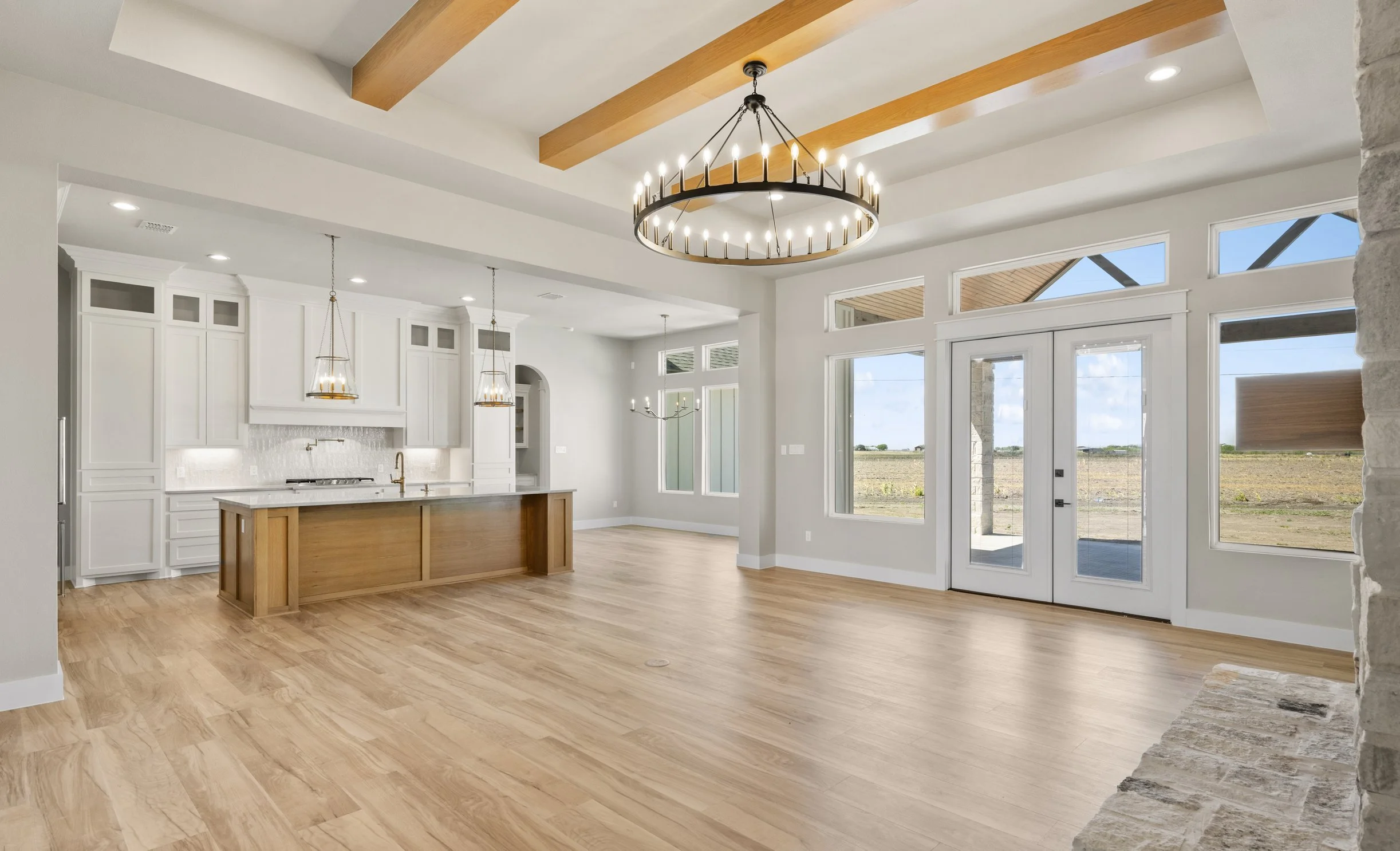 Open-concept living space with white kitchen cabinets, a wooden kitchen island, hardwood flooring, large windows, and modern lighting fixtures including a chandelier and pendant lights. Outside view shows a field under a blue sky.