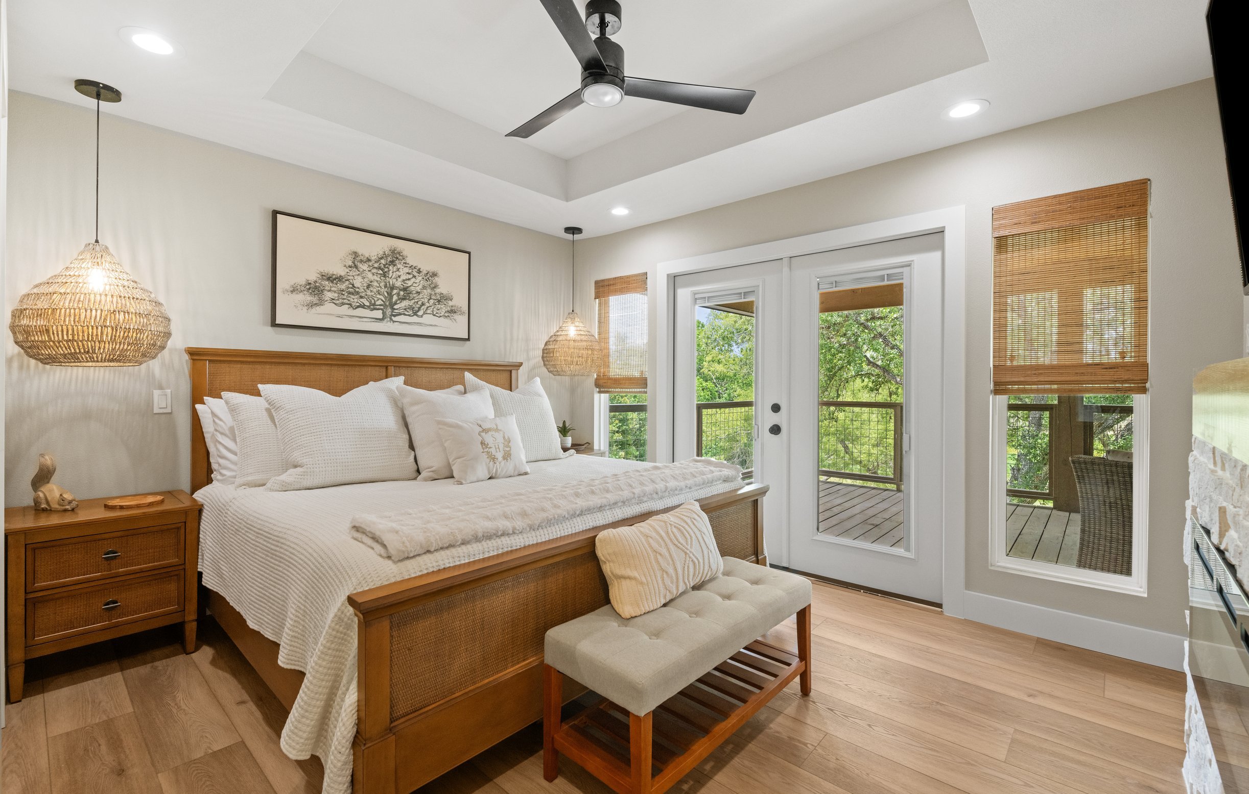 A cozy bedroom with a wooden bed frame, white bedding, and decorative pillows. Two woven pendant lights hang above matching nightstands, and a framed tree picture decorates the wall. Large windows and a glass door lead to a balcony with greenery outs