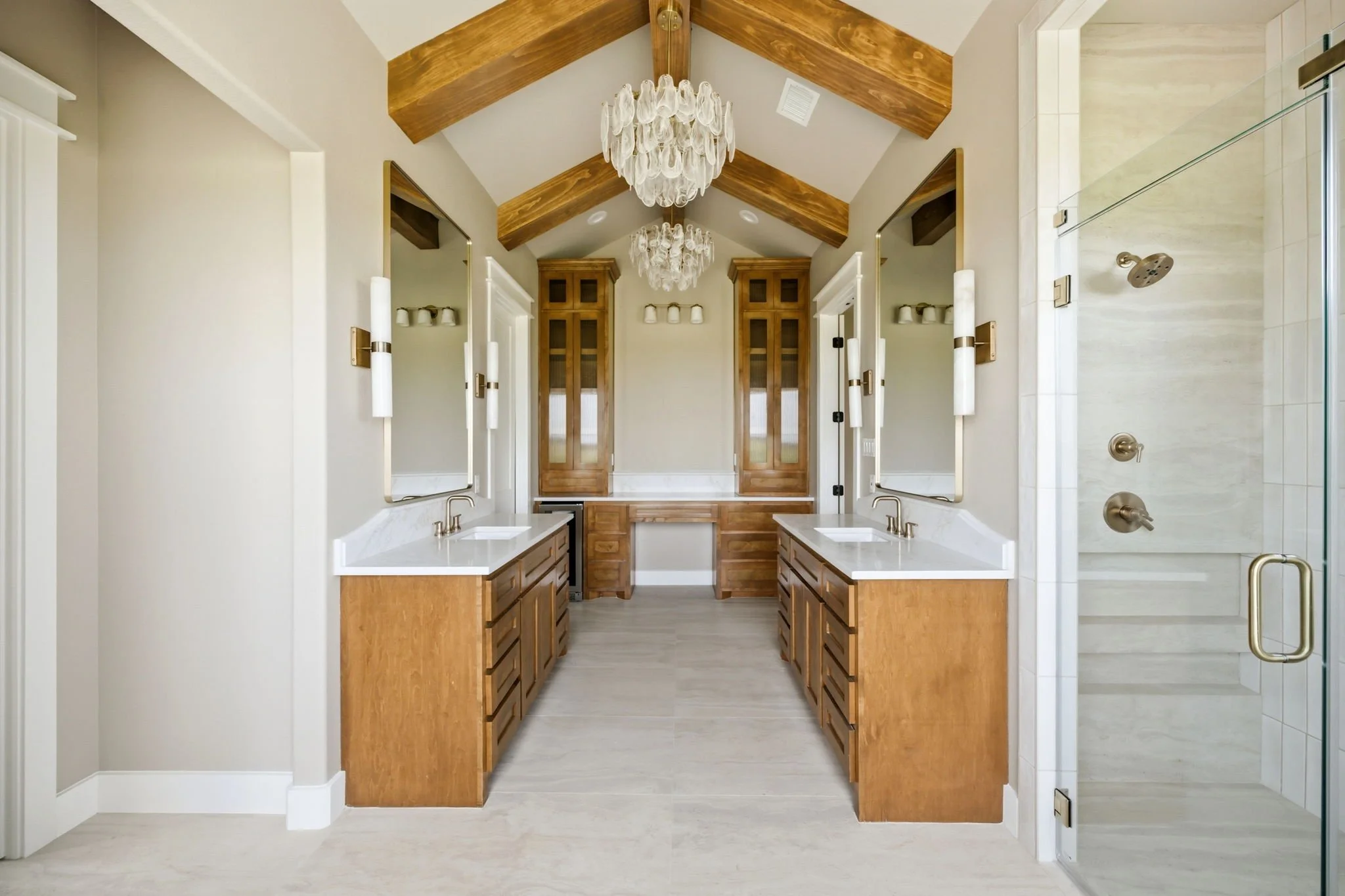 A luxurious bathroom with double vanity sinks, wooden cabinets, large mirrors, and a glass-enclosed shower with a brass showerhead. The ceiling has exposed wooden beams and two elaborate chandeliers hanging in the center.