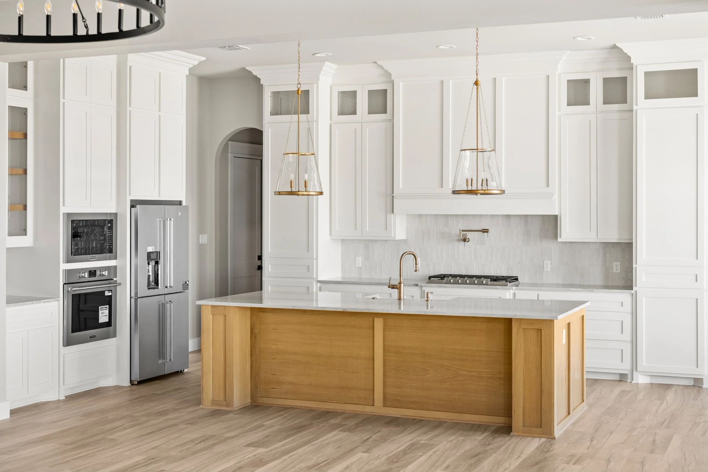 Modern white kitchen featuring tall cabinetry, a central island with wood finish, double ovens, a refrigerator, and pendant lights above the island.