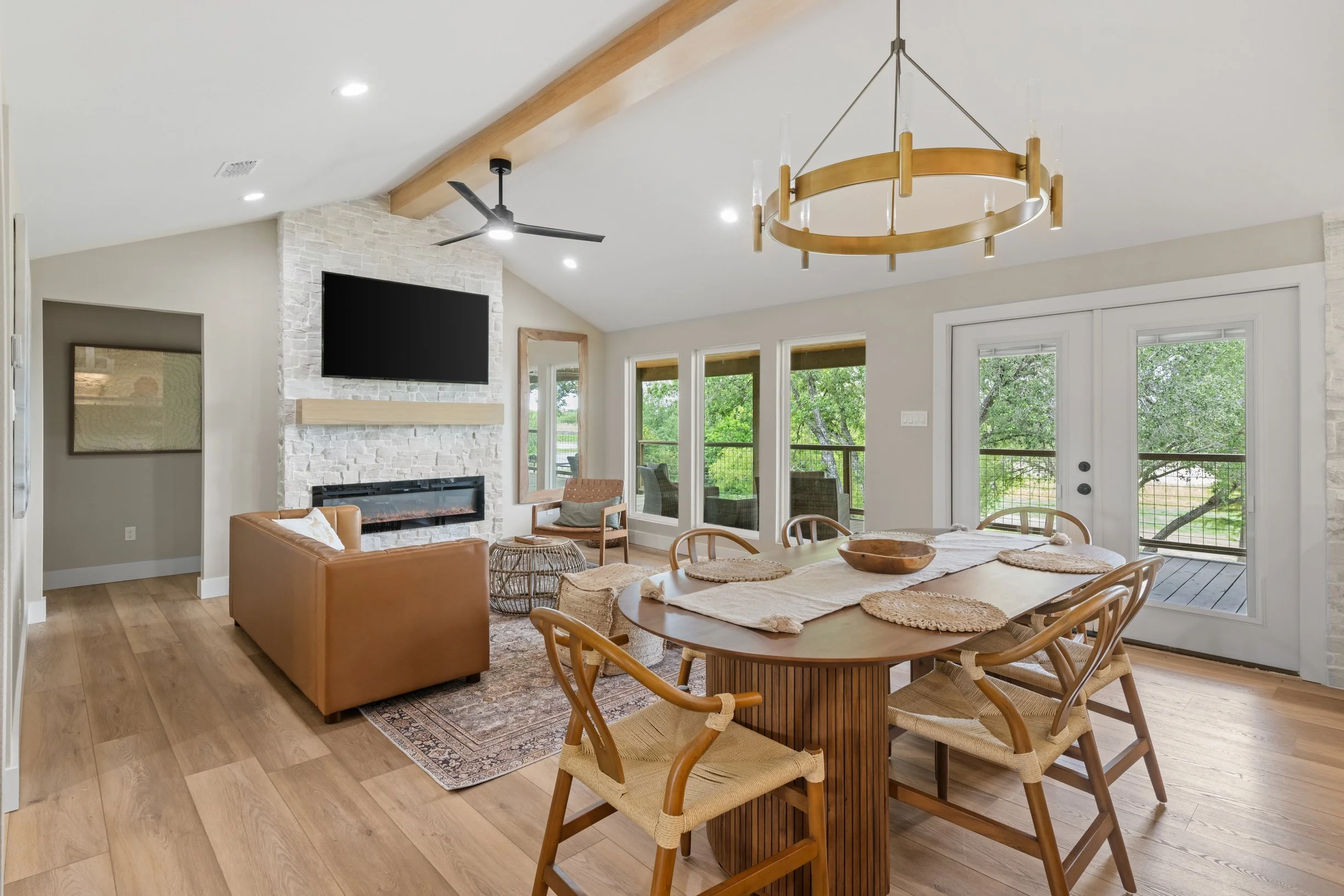 Open-concept living and dining area with a white stone fireplace, wall-mounted TV, wooden ceiling beams, large sliding glass doors, and a round dining table surrounded by wooden chairs.