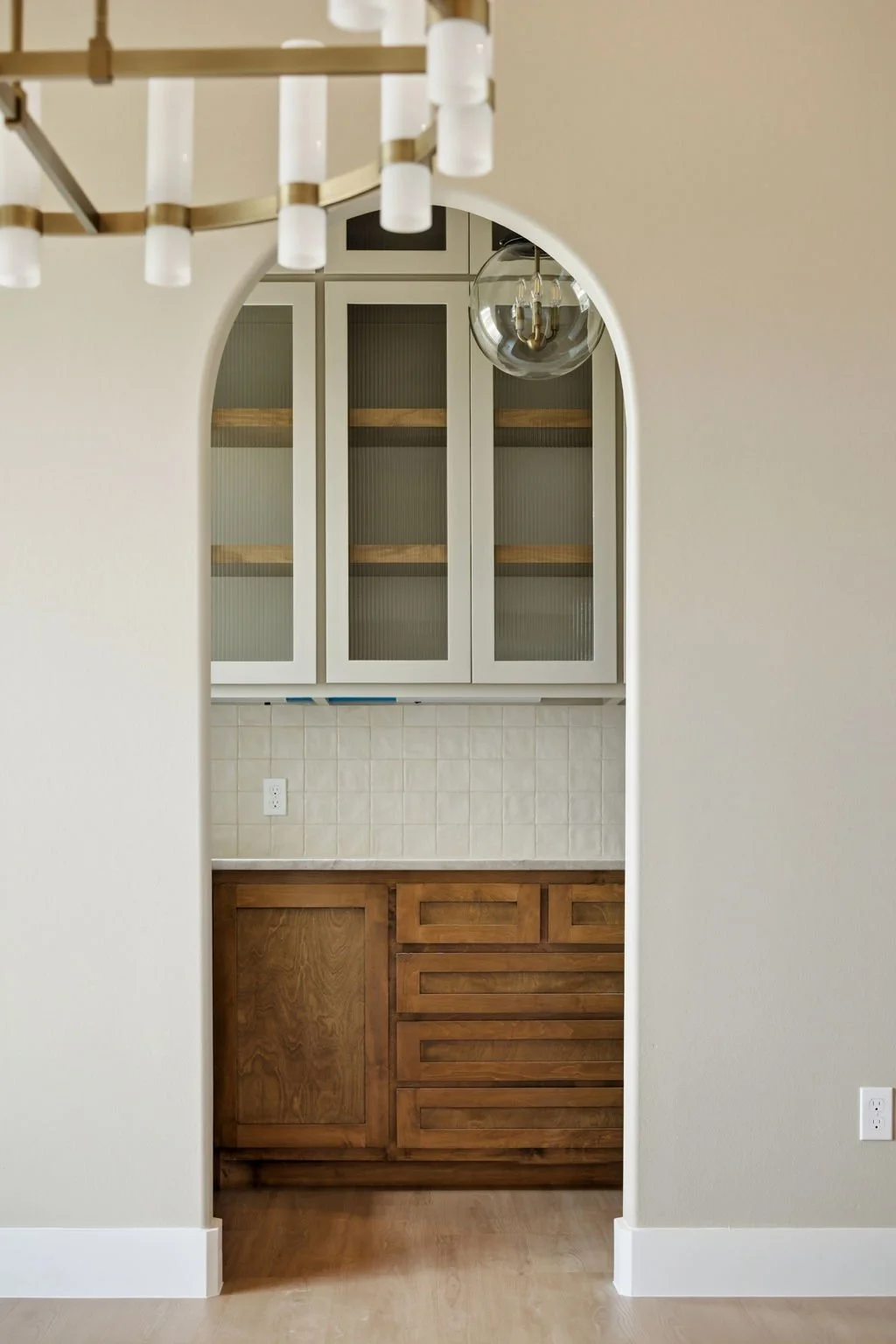 Kitchen area seen through an archway with wooden lower cabinets, glass cabinet doors, and a black chandelier.
