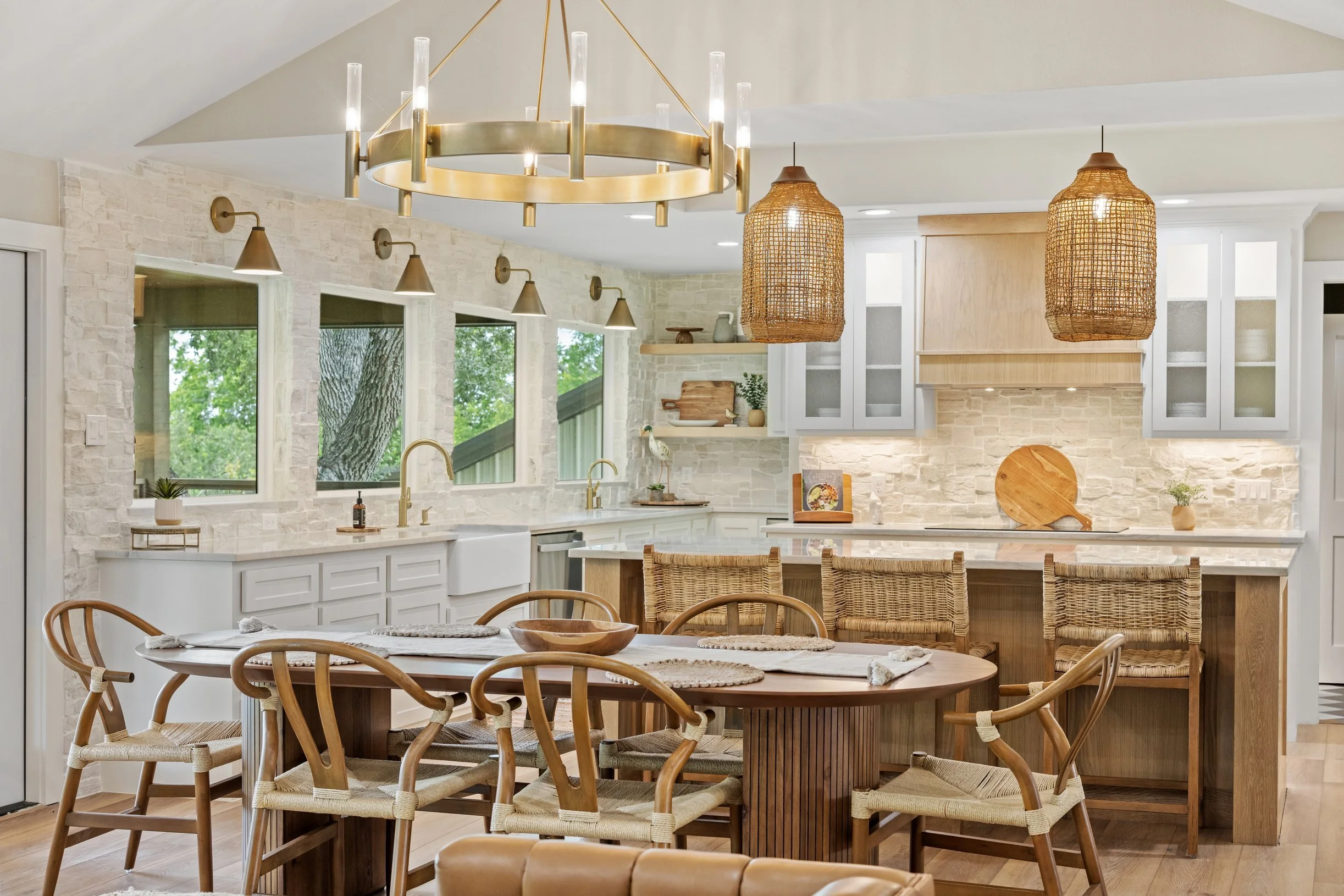 Modern kitchen with white cabinets, stone walls, and wooden furniture, featuring a round dining table with woven chairs and distinctive pendant lights.