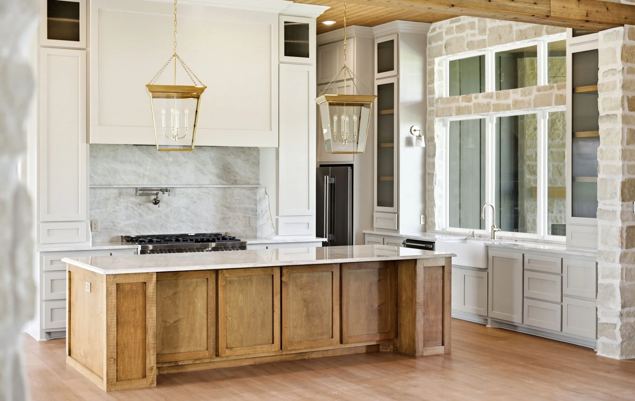 Modern kitchen with white cabinetry, a marble backsplash, and a large island with a wood finish. There are two gold pendant lights hanging above the island. The kitchen features a stone wall and large windows letting in natural light, with a view of 