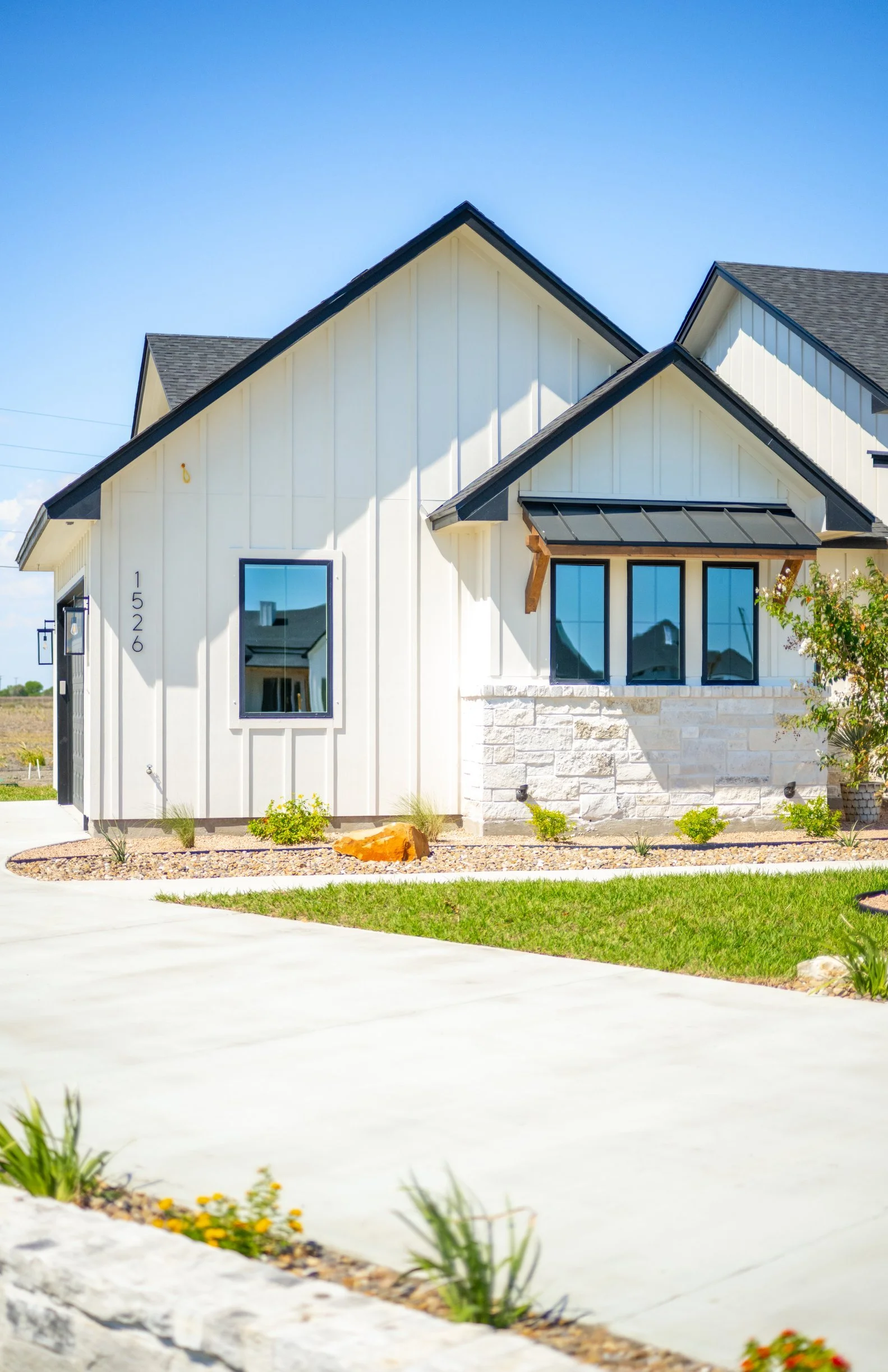 Modern house with white exterior, black trim, large front window, and a small attached roofed porch, surrounded by landscaped yard with rocks, small plants, and a grassy area, under a clear blue sky.
