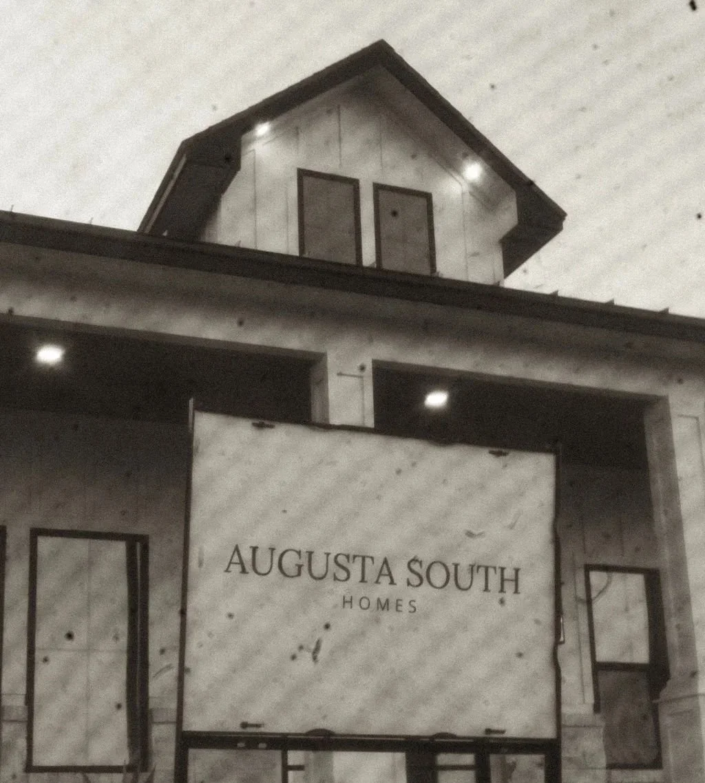 A sign reads 'Augusta South Homes' in front of a modern house with a dark roof and large windows, under a gray sky.