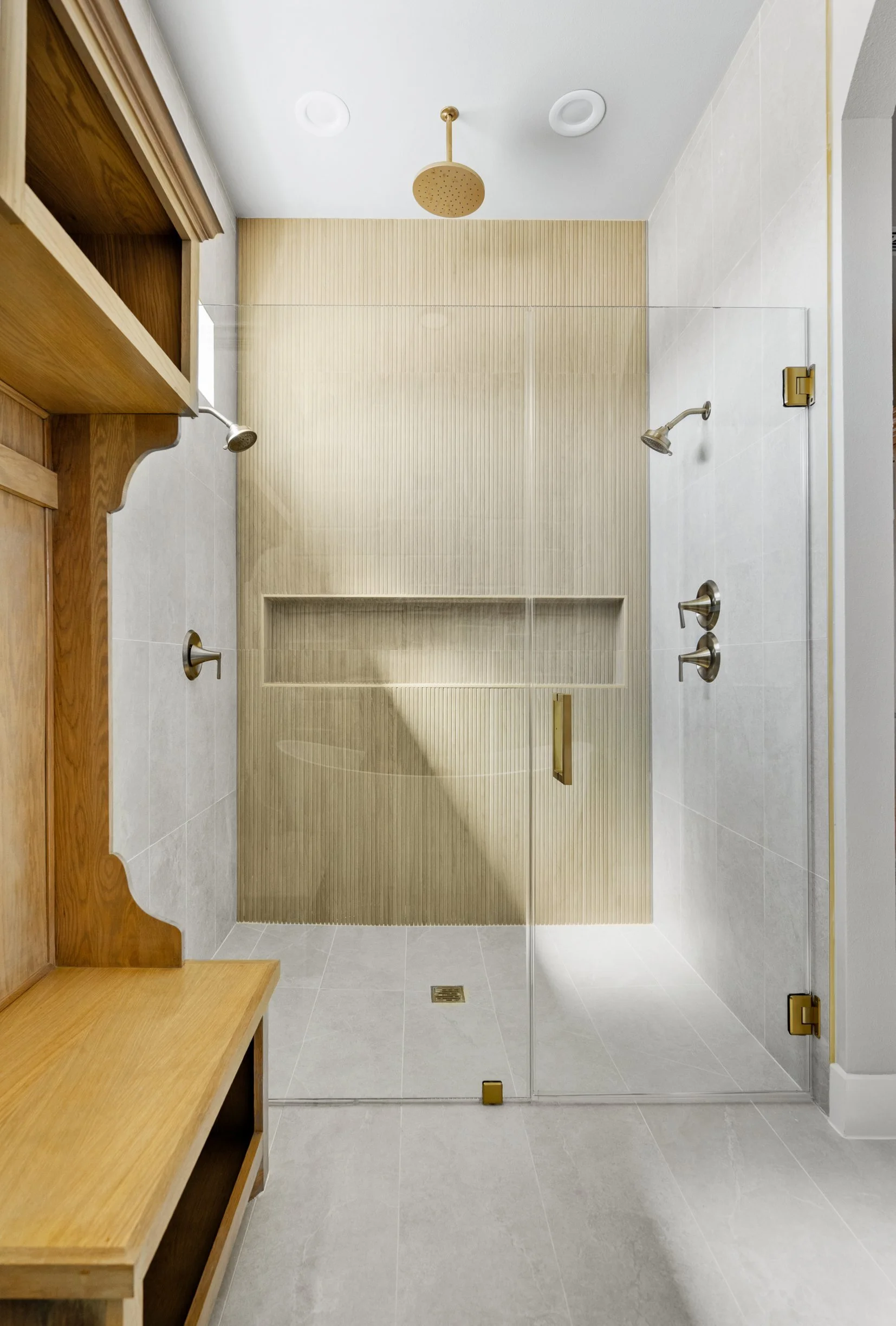 Modern walk-in shower with glass door, beige tiled walls, and two showerheads, one on each side. There is a niche in the tiled wall and a round showerhead on the ceiling. The floor is tiled in light gray.