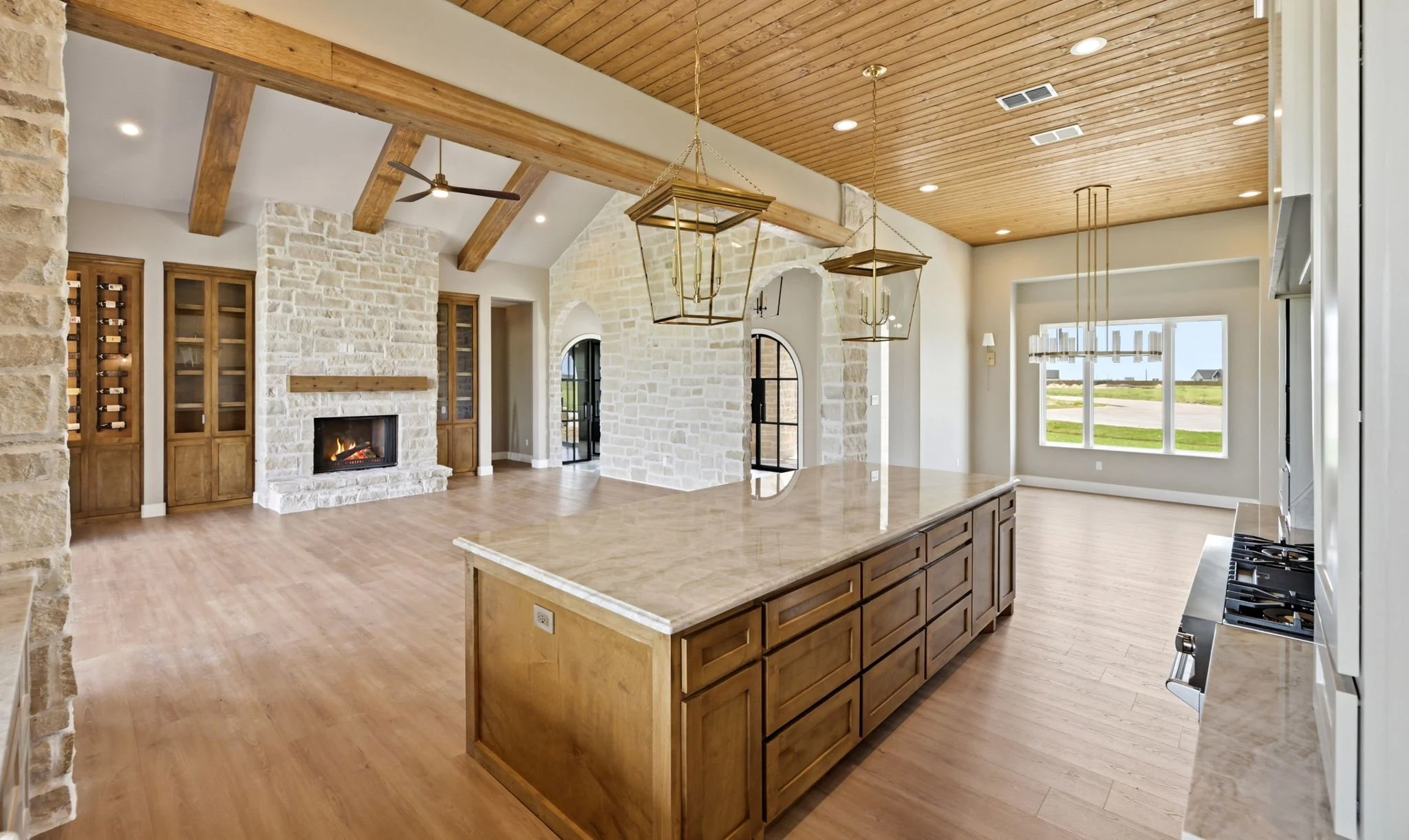 Interior view of a spacious kitchen and living area with wooden ceiling, exposed beams, a stone fireplace, and large windows showing an outdoor landscape.