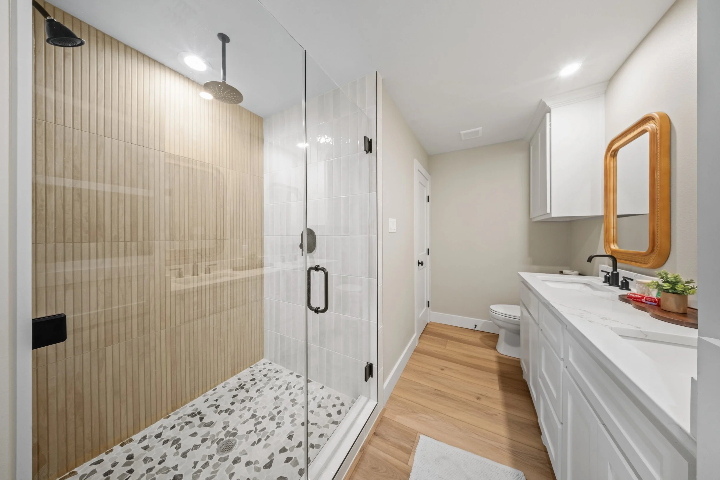 A bathroom with a walk-in shower with a glass door, beige wood-like tile wall, terrazzo floor, white vanity with marble countertop, black fixtures, a mirror with a wooden frame, and a toilet.