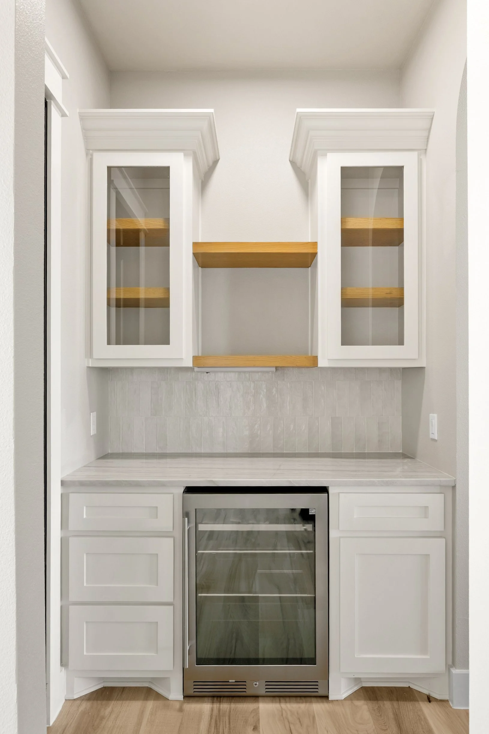 White kitchen cabinetry with glass doors, wooden shelves inside, a built-in wine cooler, and a white countertop on a hardwood floor.