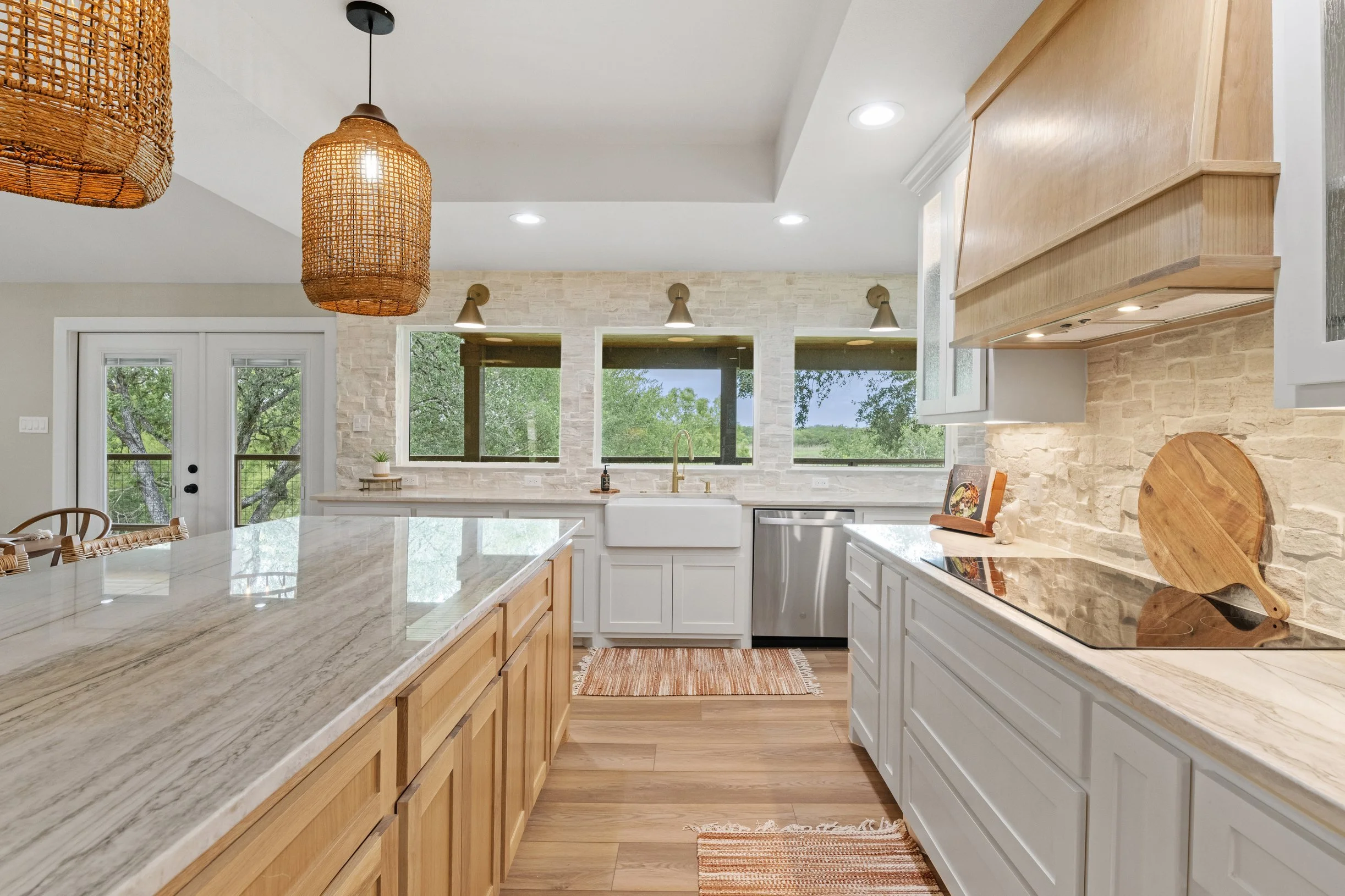 A modern kitchen featuring white cabinets, light wood accents, a marble countertop, pendant lights, and large windows showing greenery outside.