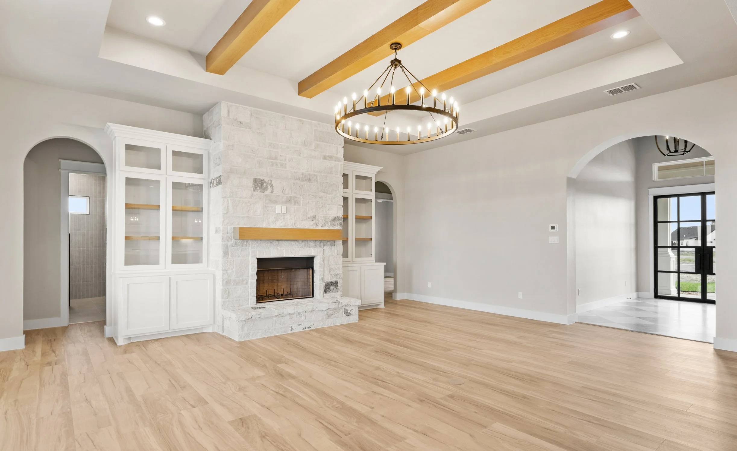 Empty living room with hardwood floors, a white brick fireplace, built-in white cabinets, a large round chandelier, and arched window and door openings.