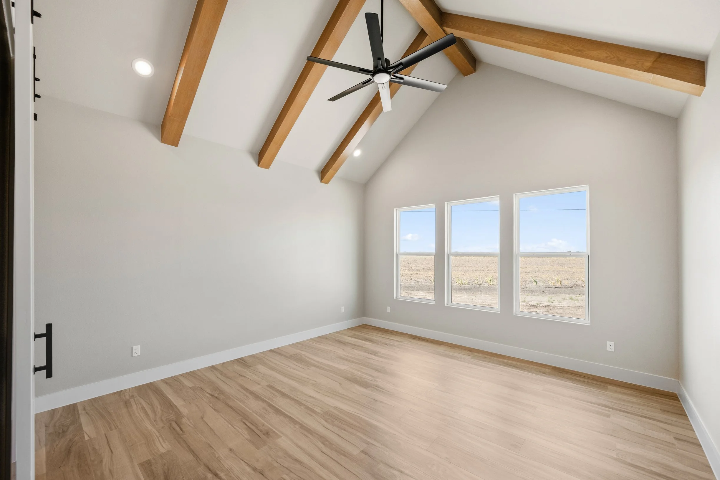 Empty room with white walls, wooden floor, large windows showing a flat landscape, and exposed wooden ceiling beams with a ceiling fan.