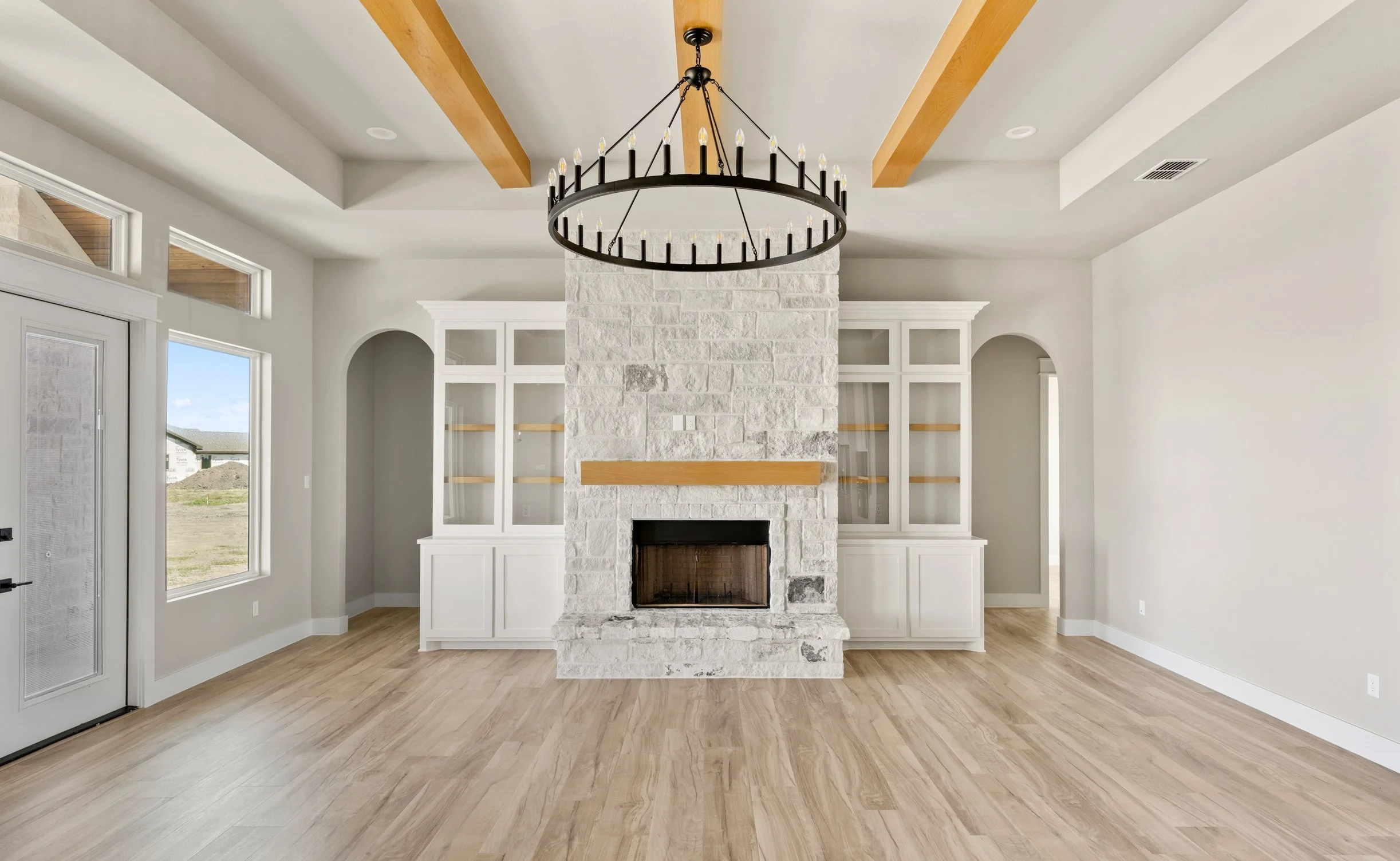 Living room with a white stone fireplace, built-in white cabinets, large windows, wooden ceiling beams, a black circular chandelier, and light wood flooring.