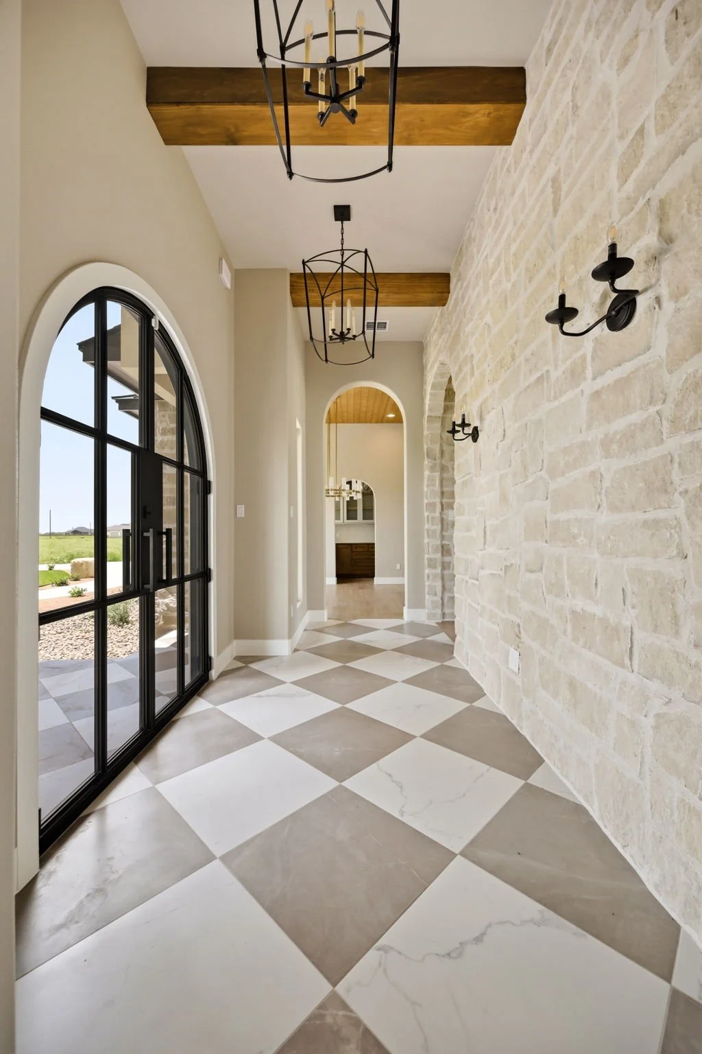 A hallway with a checkered floor, beige walls, black metal chandeliers, and a stone accent wall with black wall sconces. Large arched window with black frame on the left, and a doorway in the background leading to another room.