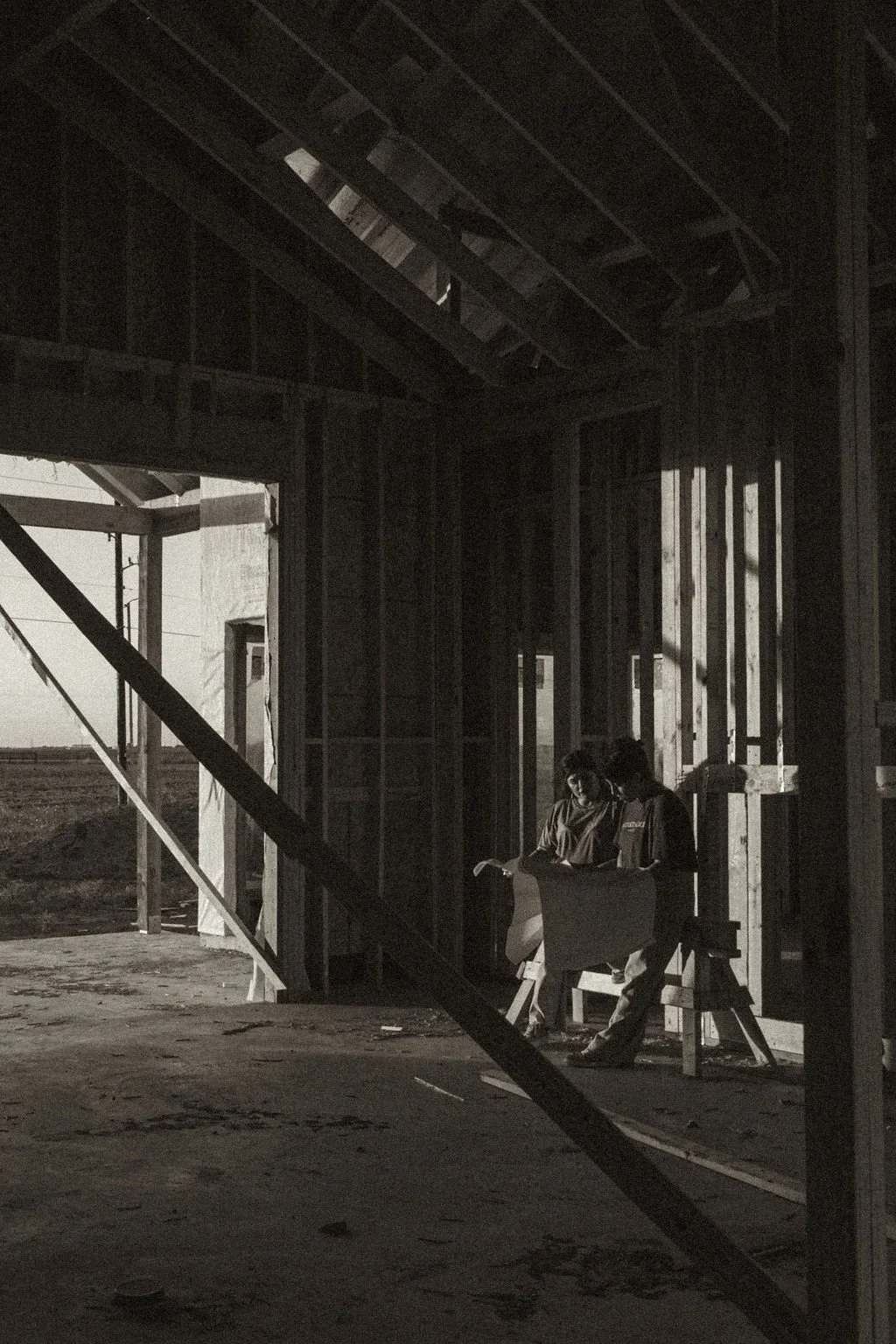 Two individuals working on a construction site inside a partially framed building during daylight, with construction materials and open wall spaces visible.