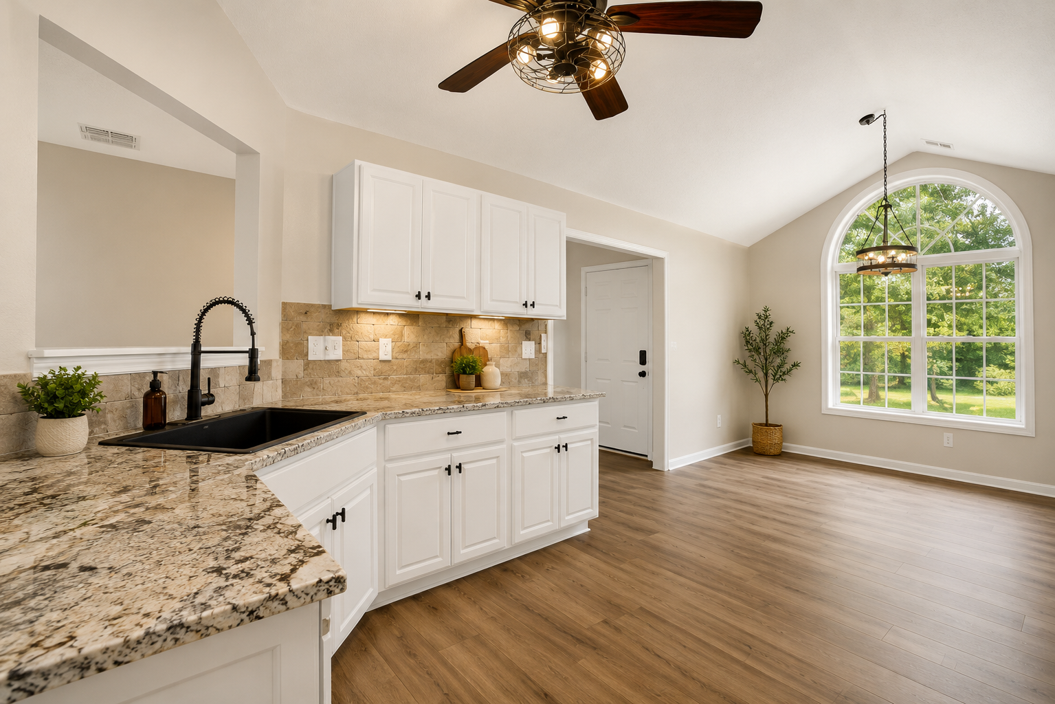 Modern kitchen with white cabinetry, granite countertops, beige tile backsplash, black sink, and black hardware. A large arched window provides a view of green trees. Wooden floors and a ceiling fan with lights are shown. Decorative plants and lighting fixtures are present.