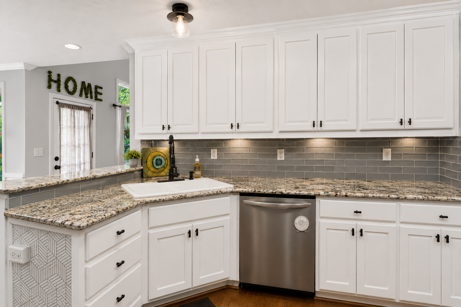 Modern kitchen with white cabinets, granite countertops, gray subway tile backsplash, stainless steel dishwasher, and a black matte faucet. A small potted plant and artwork are on the counter by the sink. Completed by Sawdust.